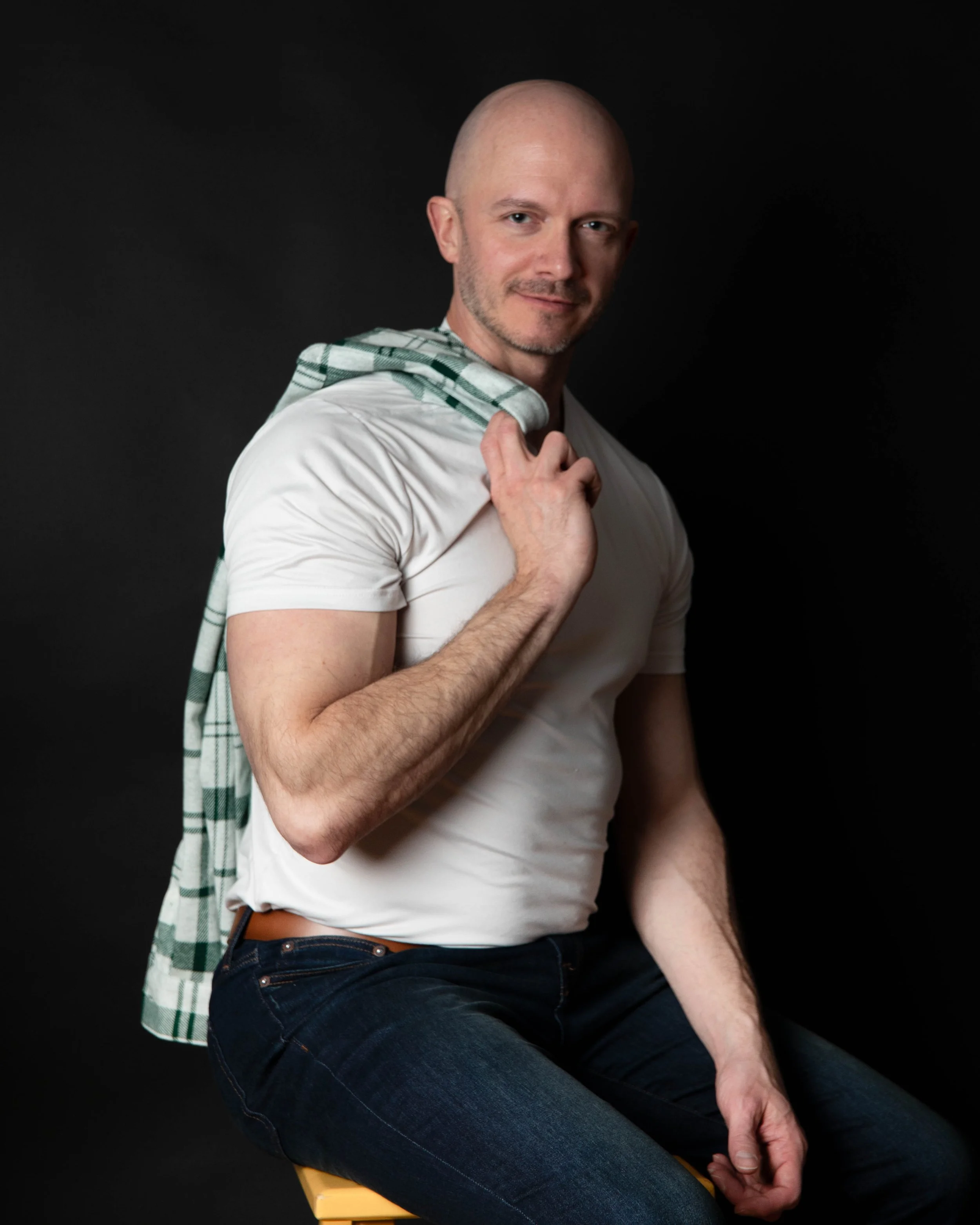 A man with a shaved head and facial hair sitting on a yellow chair, wearing a white t-shirt and dark jeans, holding a checkered shirt over his shoulder against a black background.