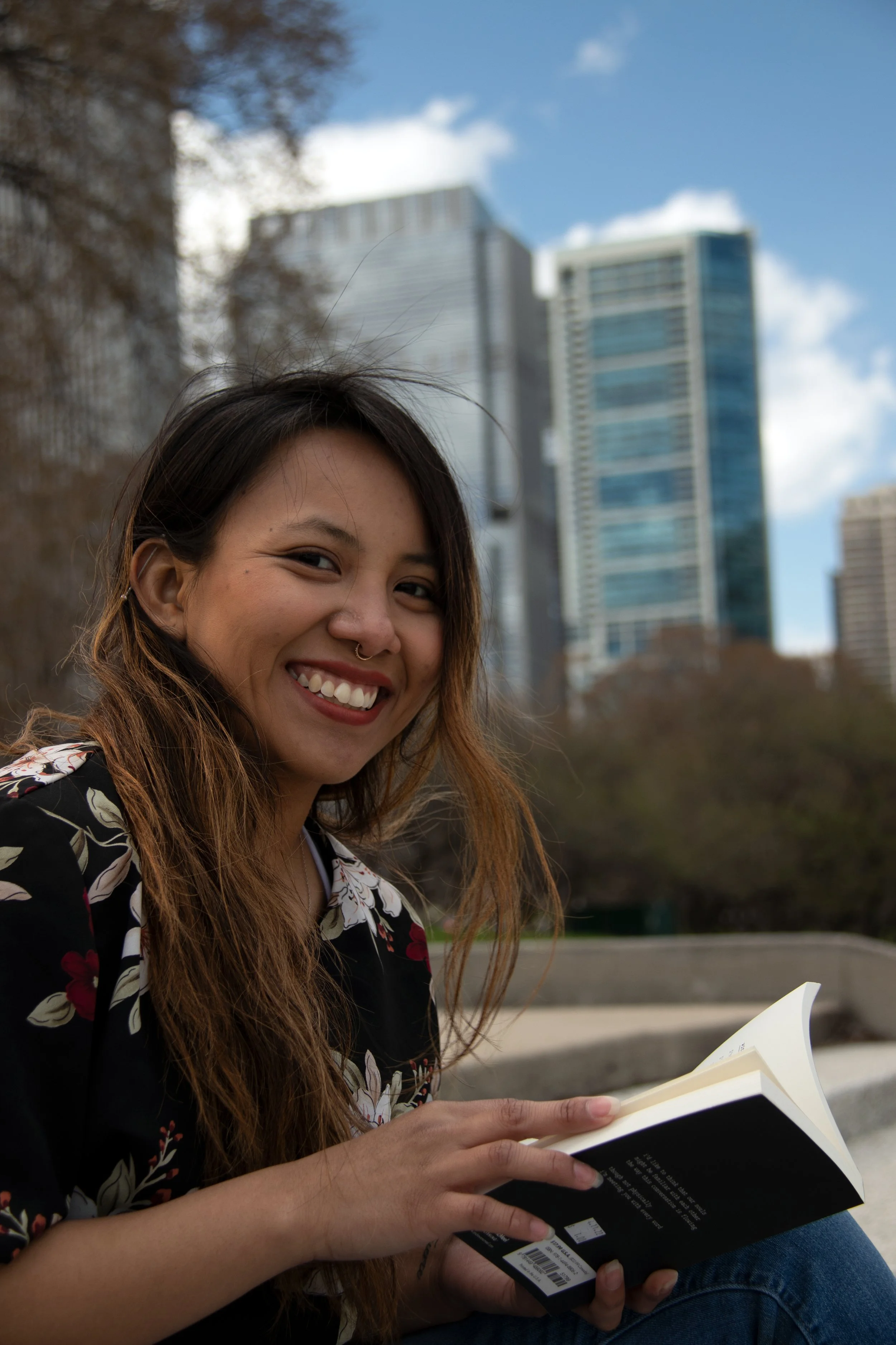 Smiling young woman with long hair, wearing a floral shirt, sitting outdoors, reading a book with buildings and sky in the background.