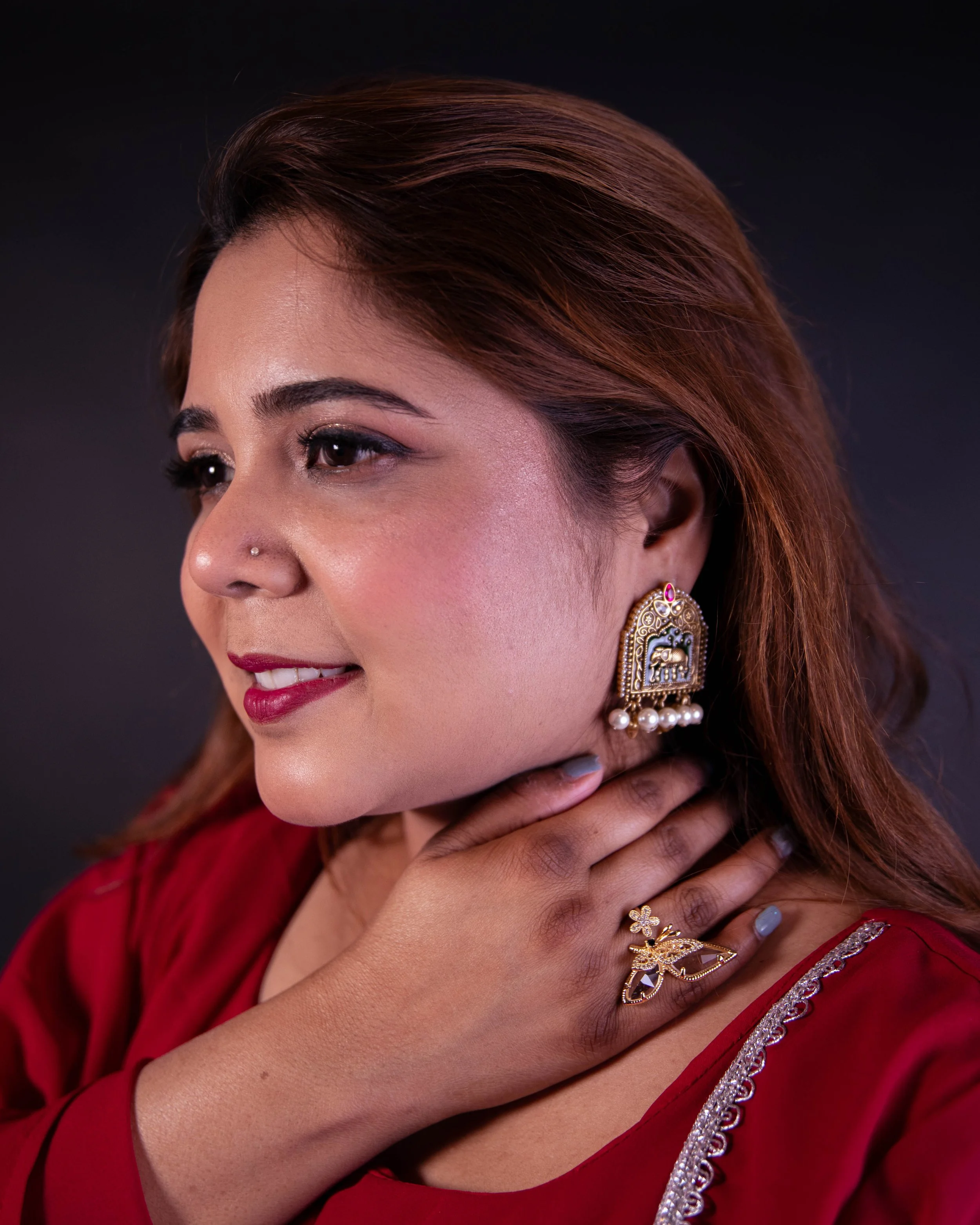 Close-up of a woman's face and hand with jewelry, wearing a red outfit with silver embroidery, against a dark background.