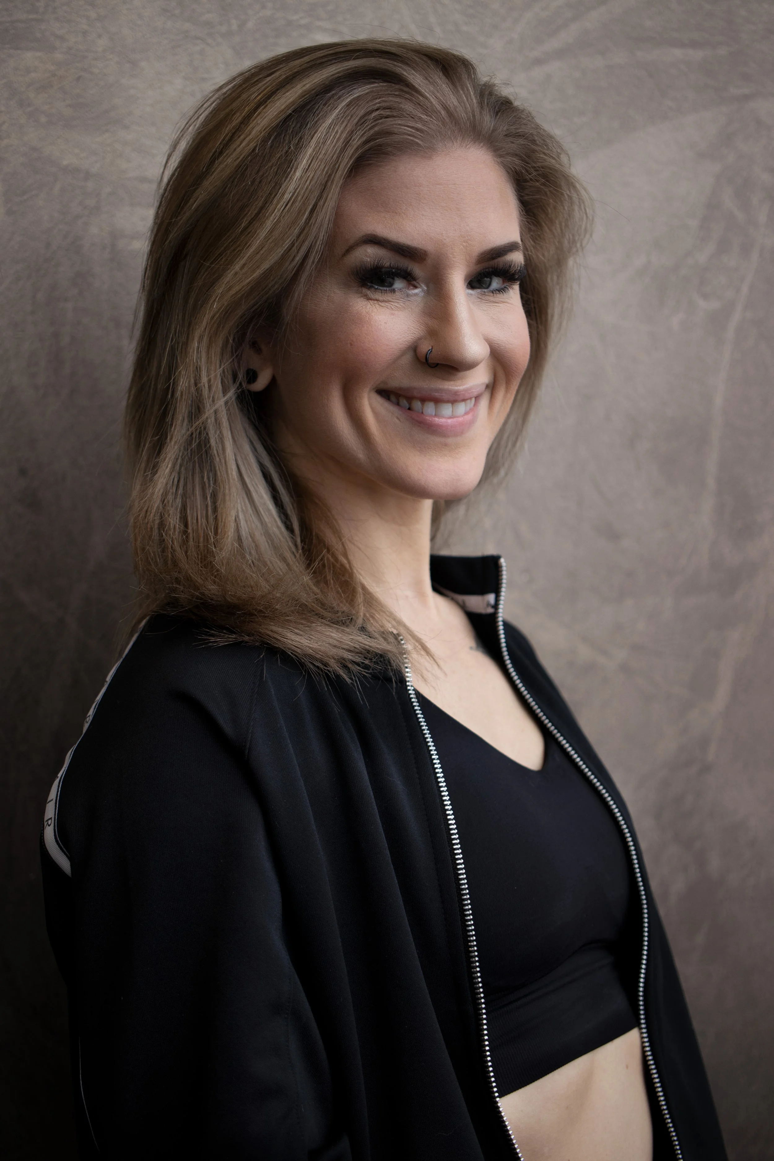 A woman with shoulder-length light brown hair, wearing a black zip-up jacket and black sports bra, smiling at the camera against a textured gray background.