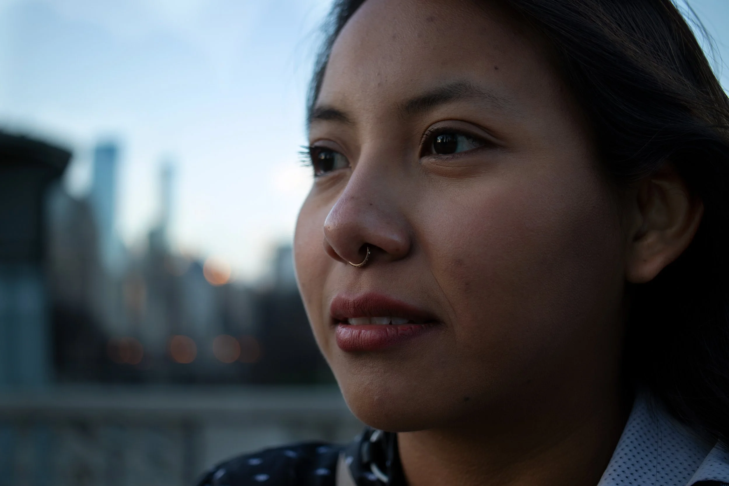 Close-up of a young woman's face with dark hair, wearing a septum piercing, looking thoughtfully into the distance against a cityscape background during the evening.