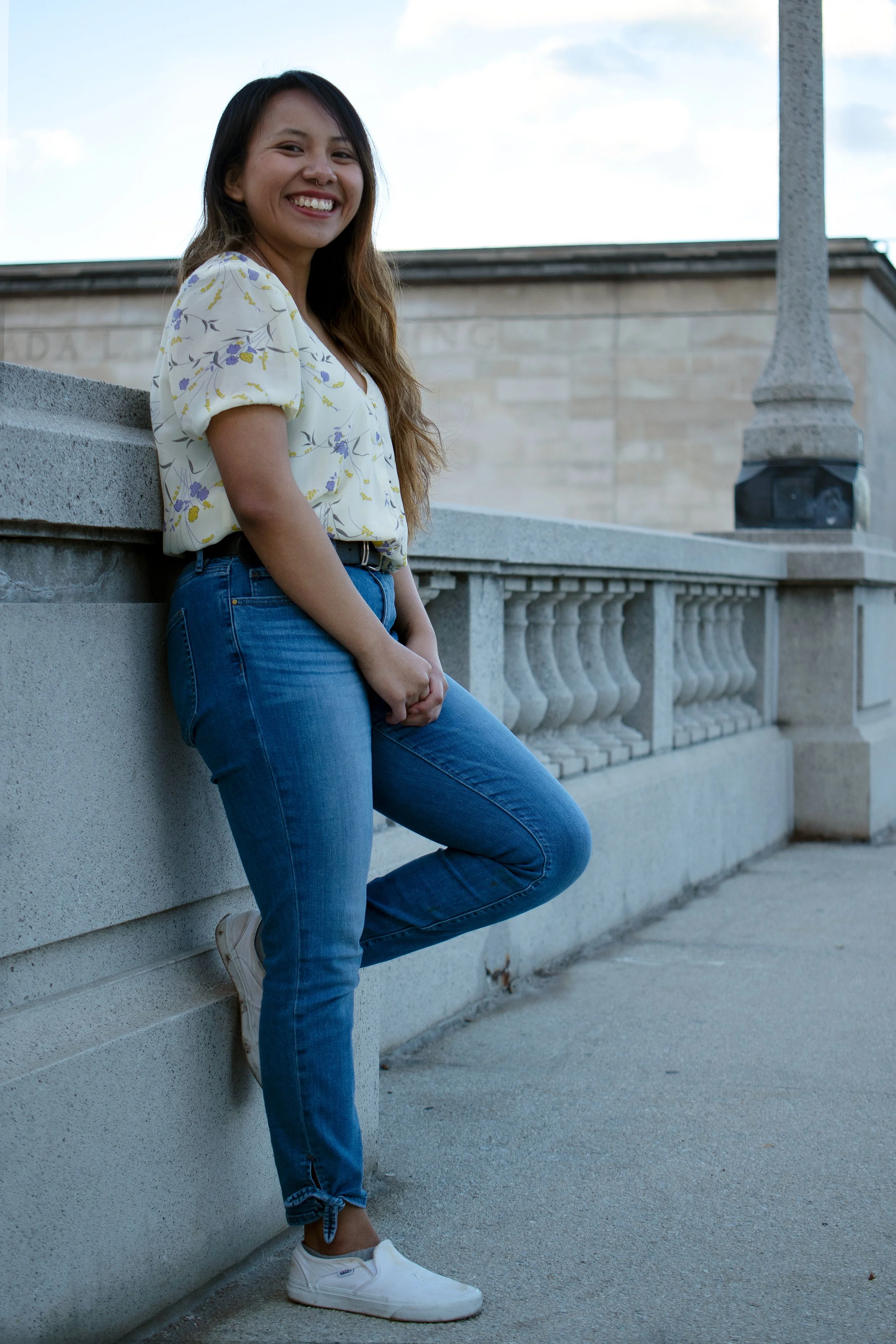 A young woman with long brown hair, wearing a floral blouse, blue jeans, and white sneakers, leaning against a stone wall outdoors with a building and a lamppost in the background, smiling at the camera.