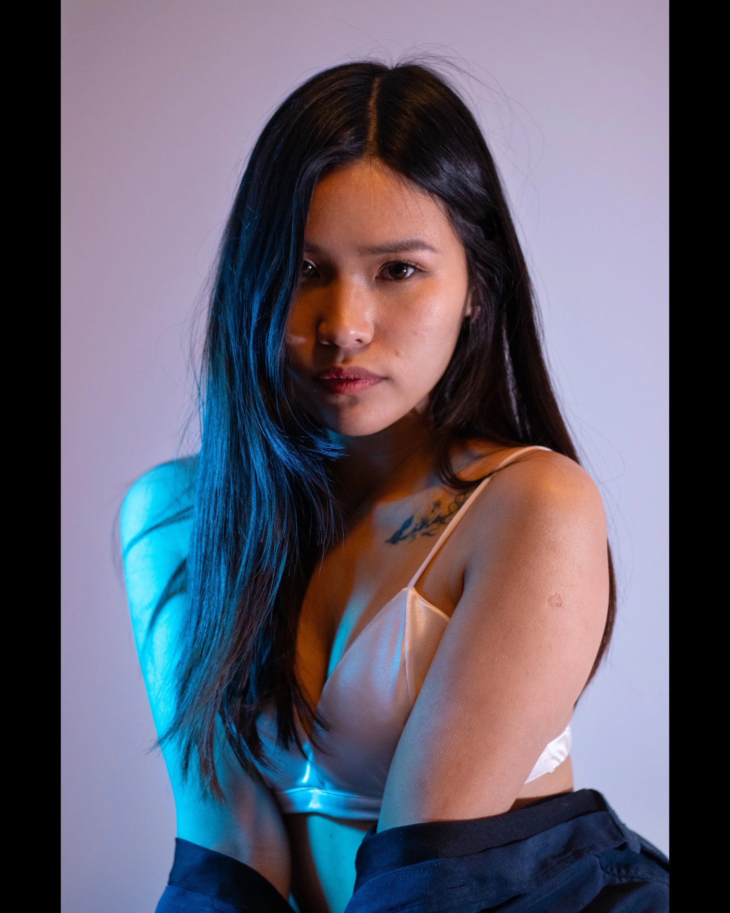 A young woman with long black hair and tan skin, wearing a white tank top and black pants, posing against a neutral background with blue and purple lighting.