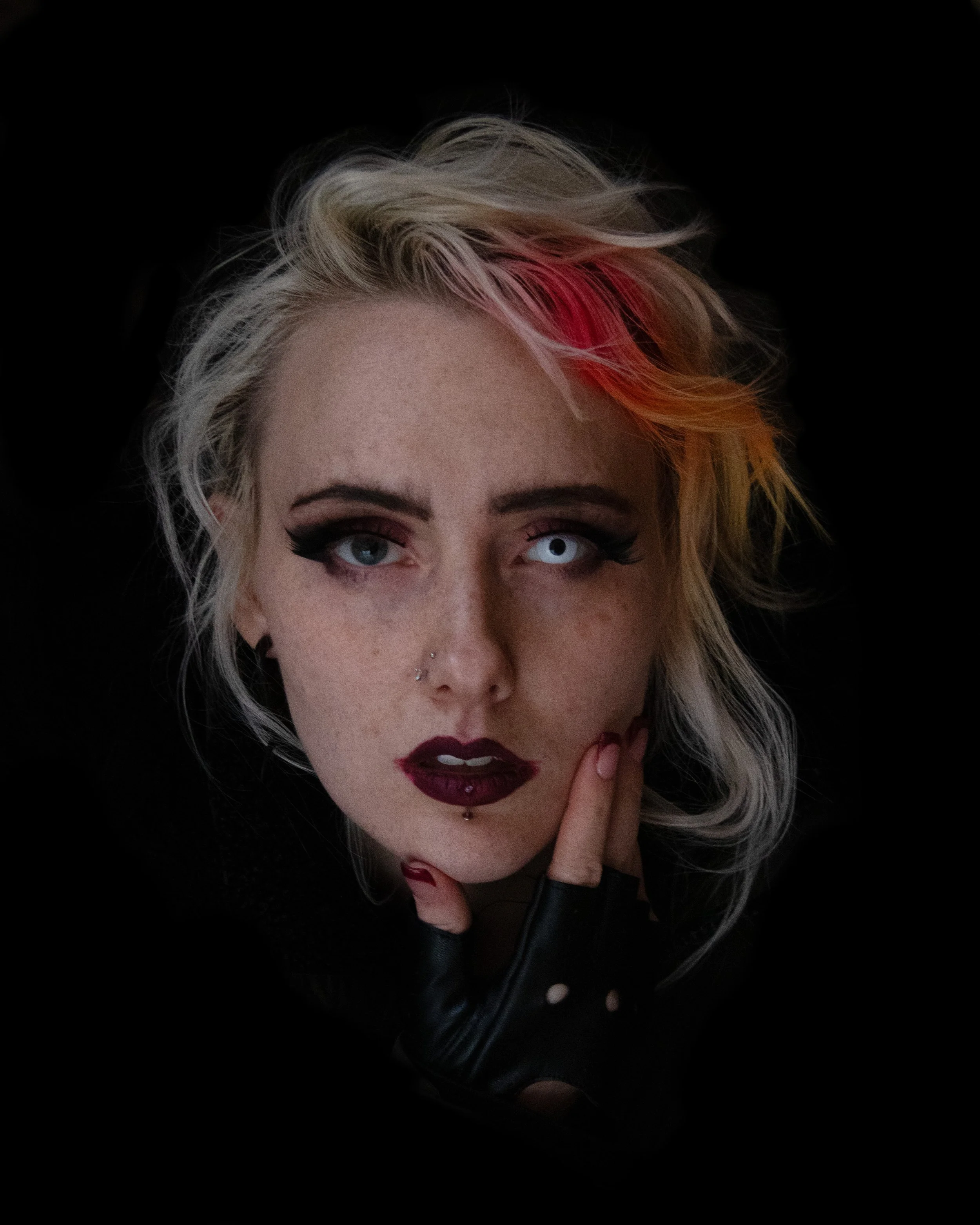 Close-up of a woman with platinum blonde, wavy hair with pink highlights, wearing dark makeup, a black leather jacket, and showing her piercing blue eyes and facial piercings, against a black background.