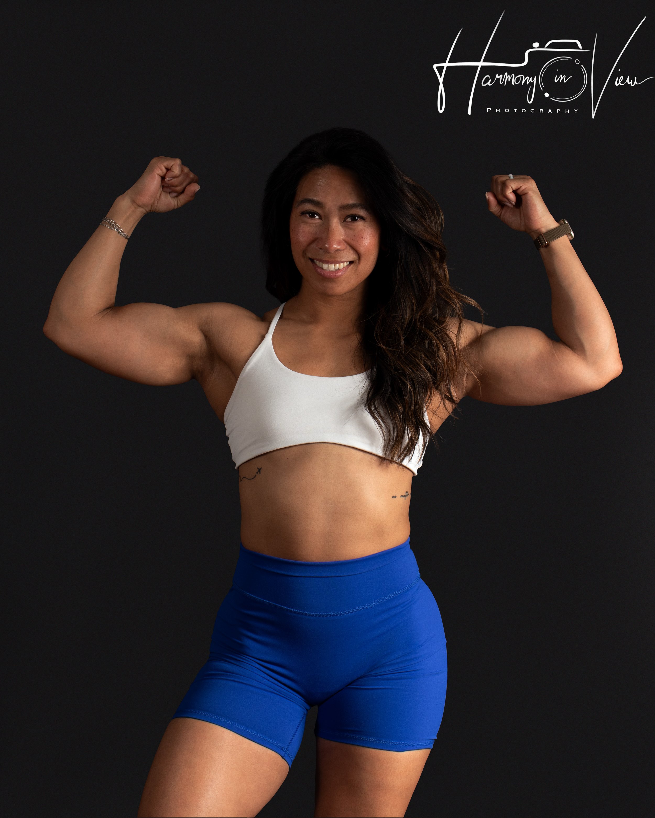 A woman with long dark hair, smiling and flexing her biceps, wearing a white sports bra and blue workout shorts, against a black background.