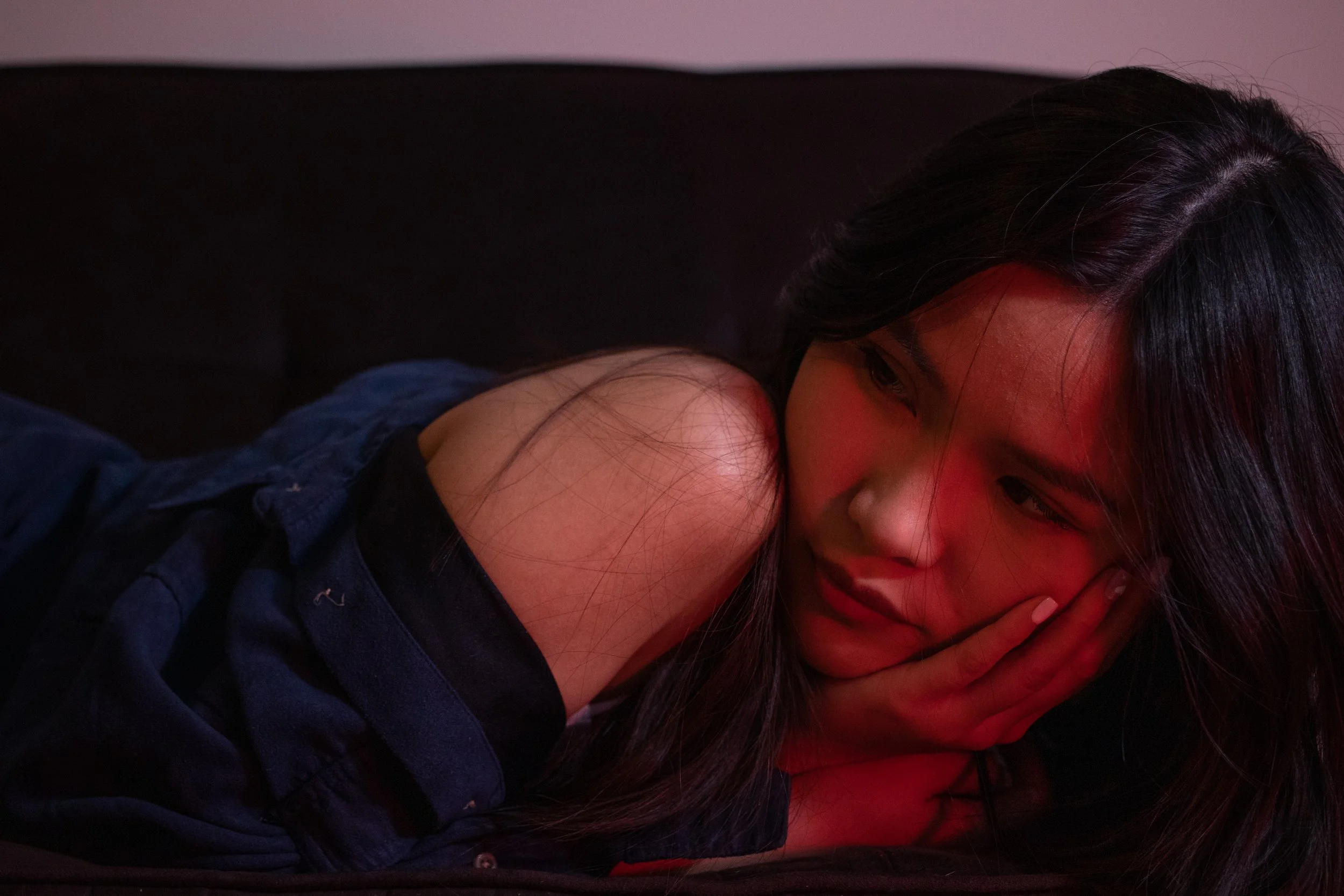 A young woman with long dark hair lying on her side, with her arm resting on her face, eyes closed, and wearing a dark jacket, in a room with dim lighting.