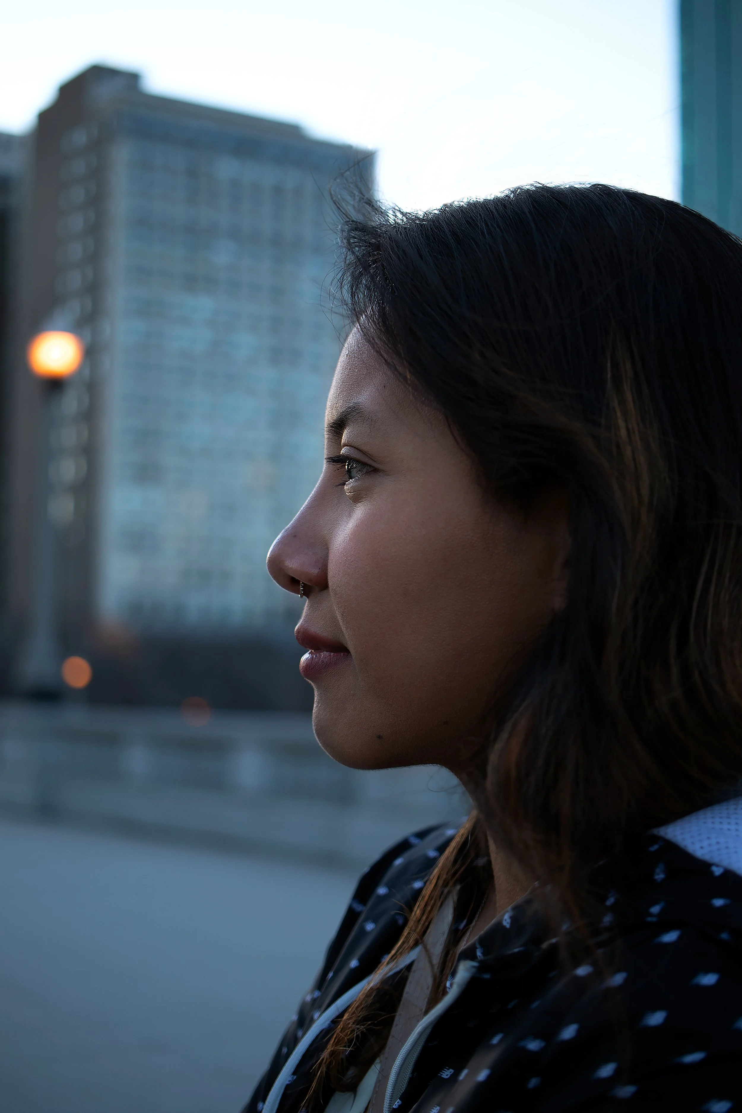 Close-up profile of a woman with medium skin tone and dark hair, standing outdoors with a cityscape and tall building in the background during dusk.