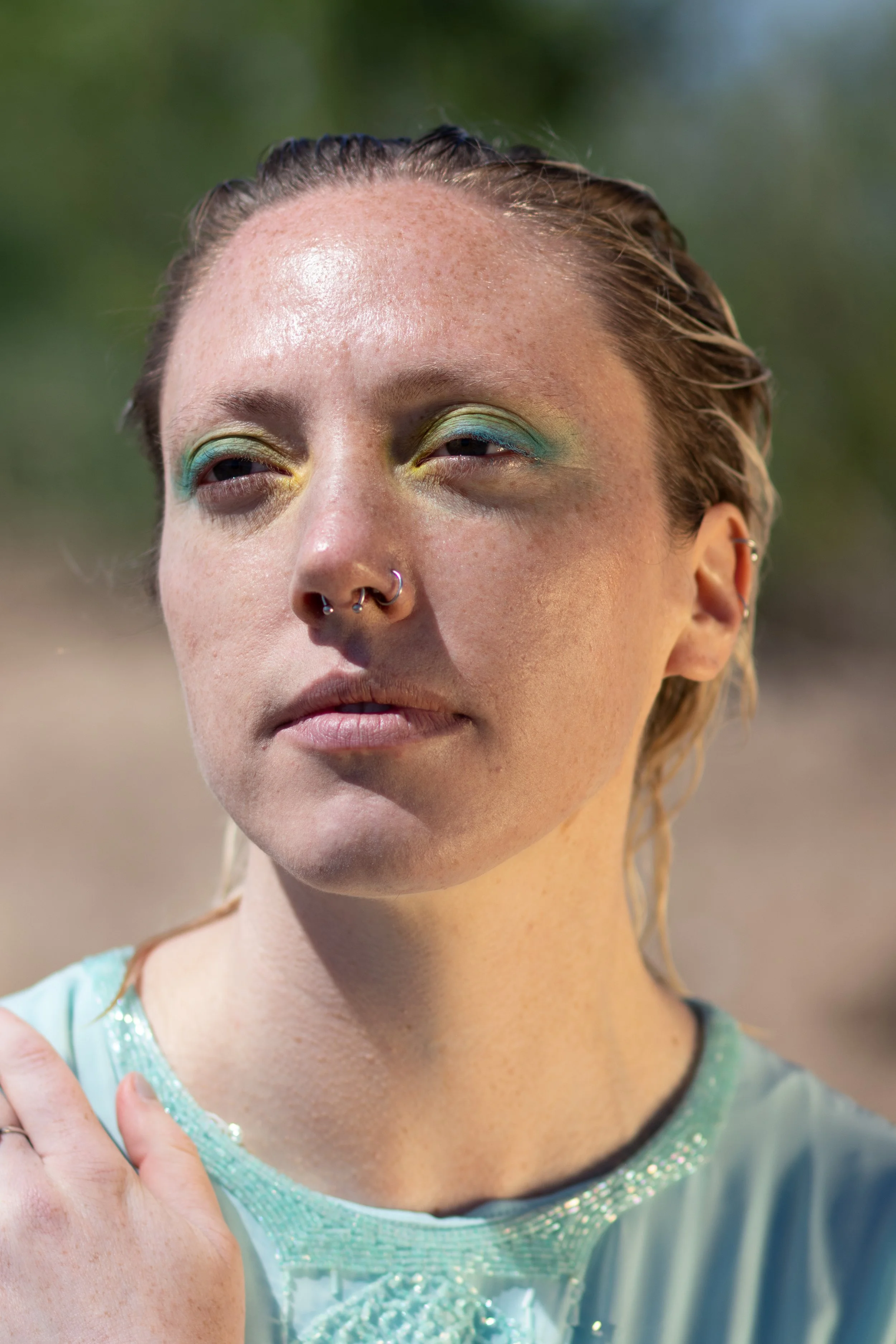 A close-up of a person with multicolored eye makeup, septum piercings, and wet hair, outdoors with a blurred background.