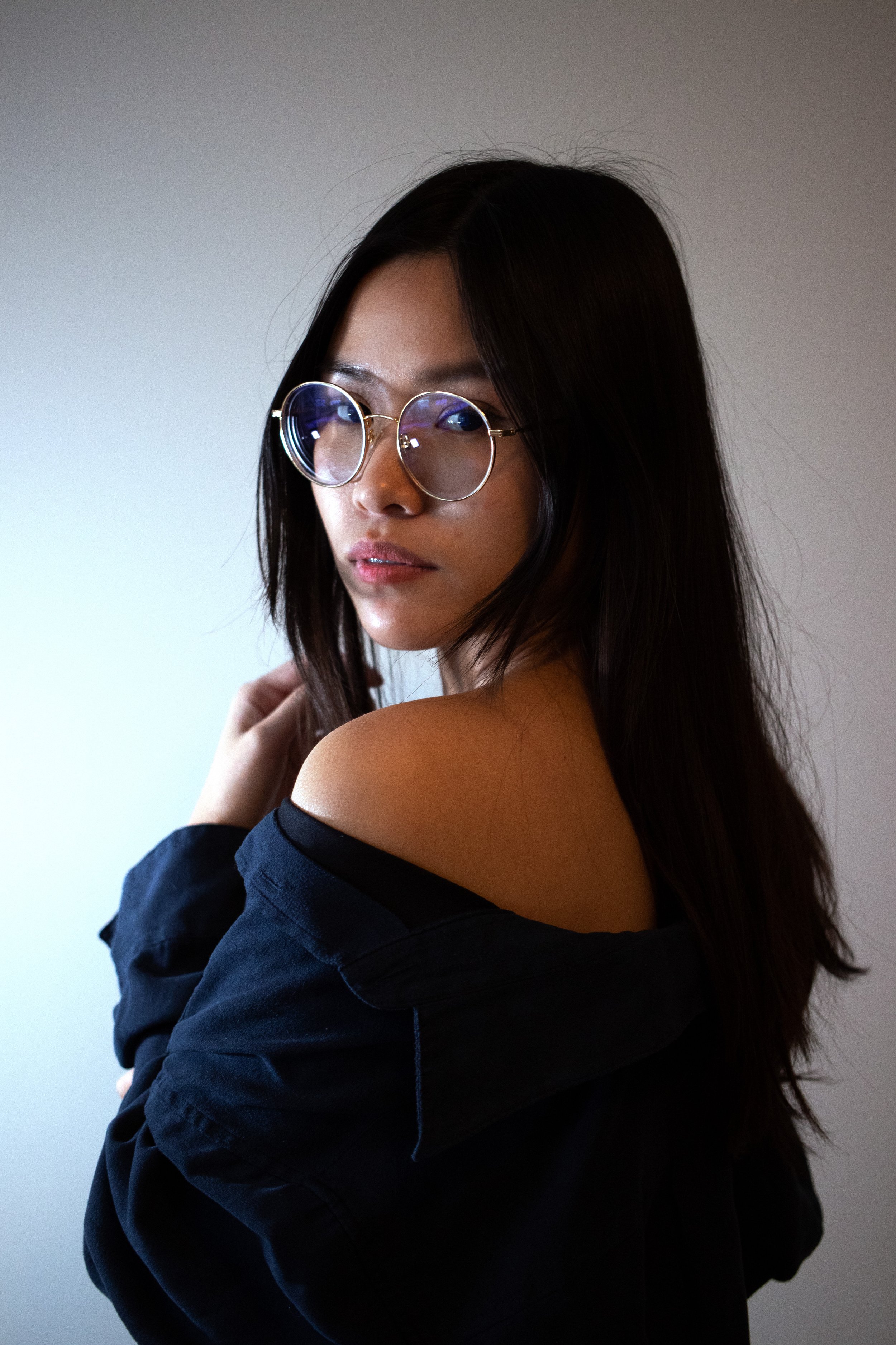 Portrait of a woman with long dark hair and glasses, wearing a black off-shoulder top, standing against a plain background.