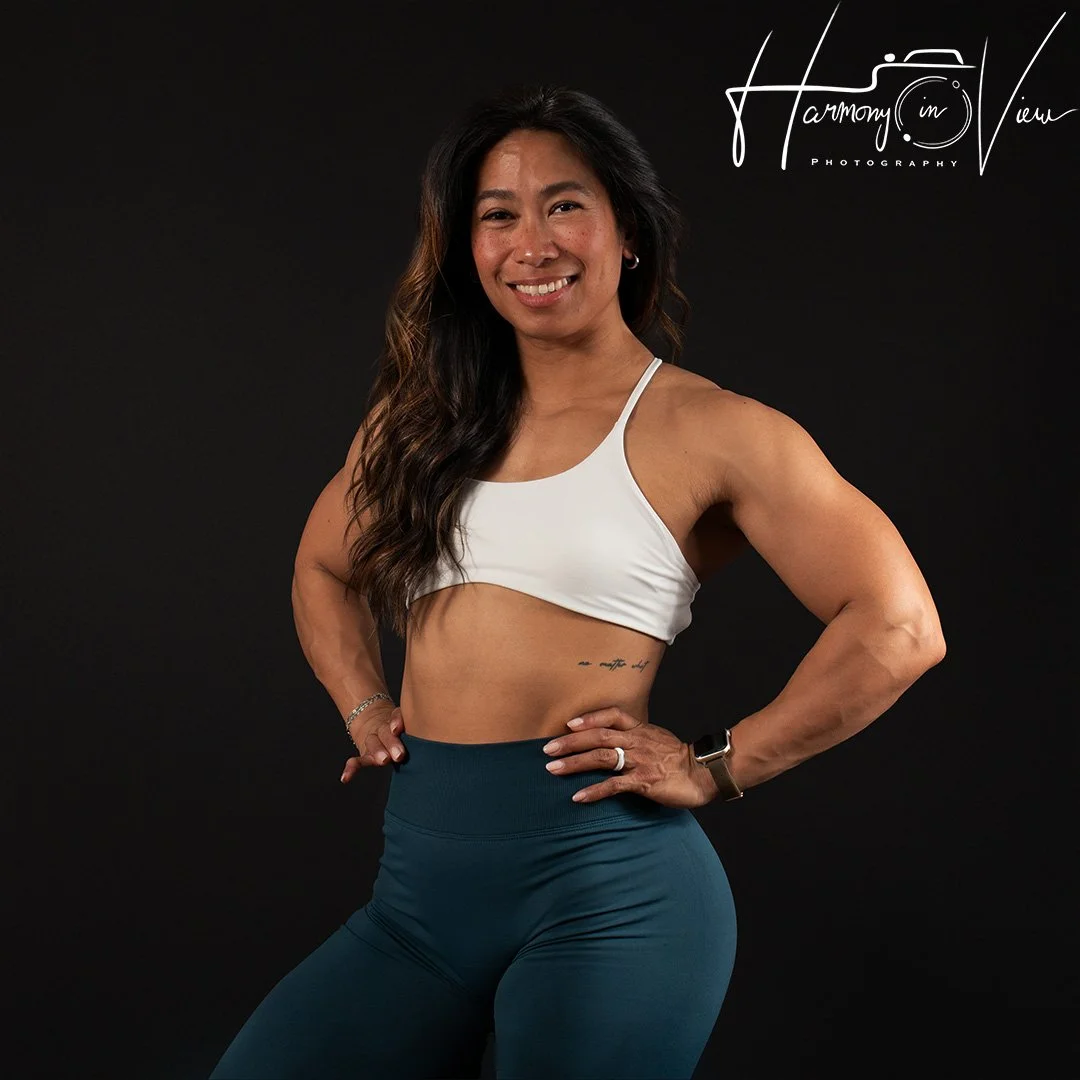 A fit woman with long dark hair, wearing a white sports bra and teal high-waisted leggings, smiling while standing against a black background.