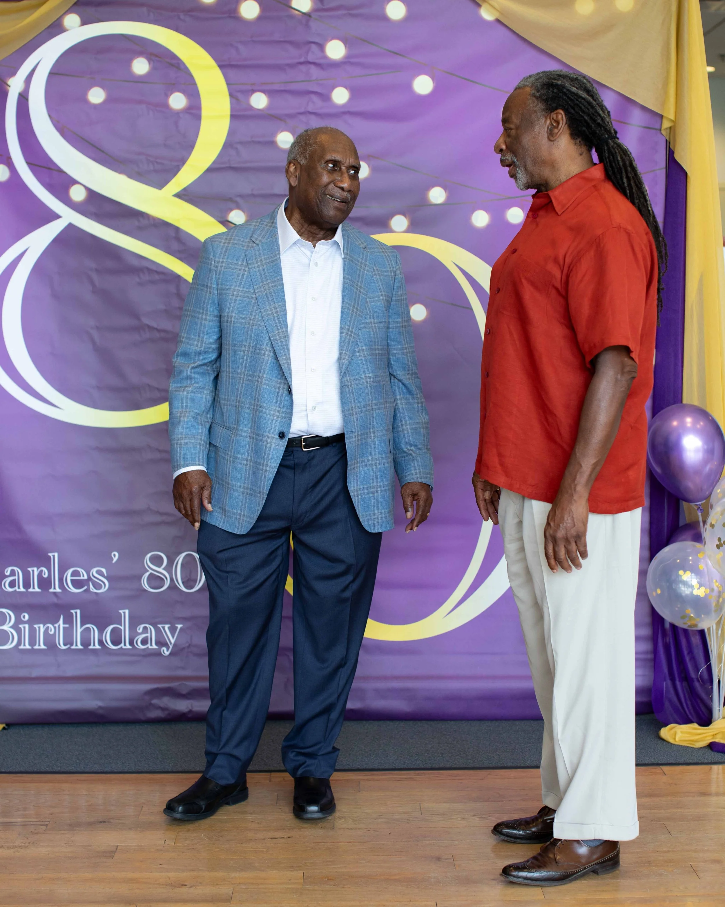 Two men in conversation at a birthday celebration, standing on a wooden floor in front of a purple backdrop with yellow and white decorations and balloons.
