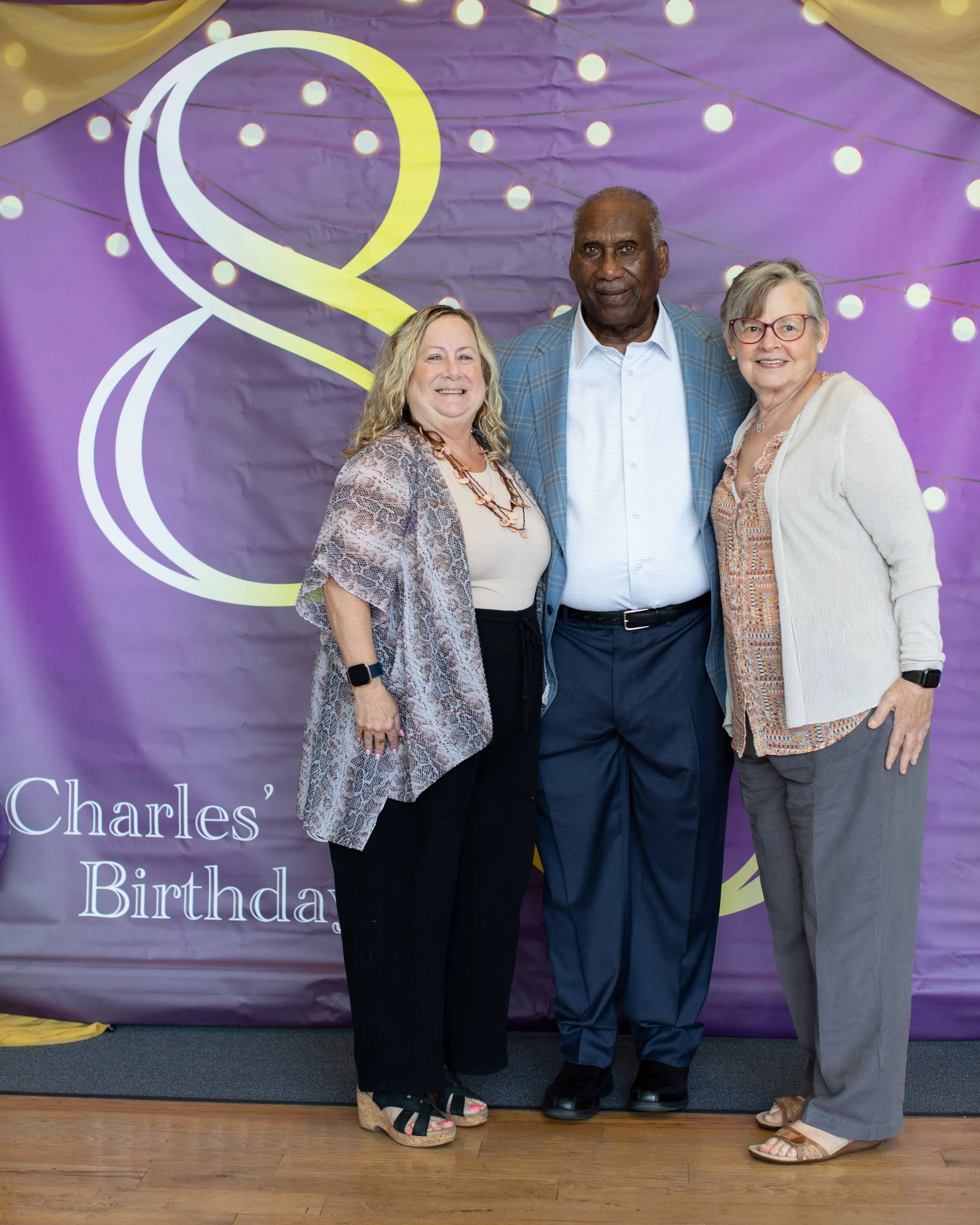 Three people standing together in front of a purple birthday backdrop with lights, smiling. Two women on either side of a man, dressed in casual formal attire, celebrating a birthday.