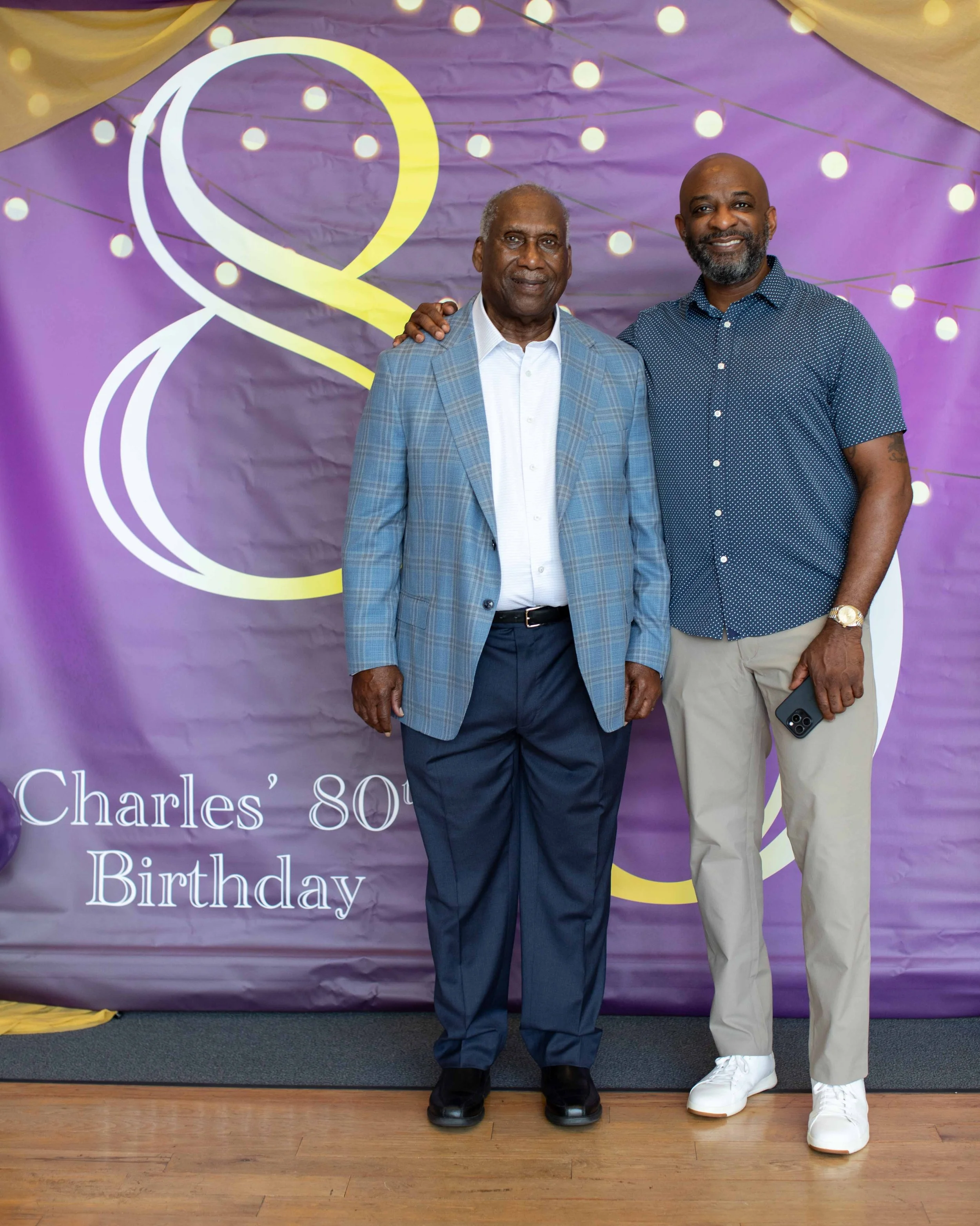Two men standing in front of a purple birthday party backdrop with the words "Charles' 80th Birthday" partially visible. The man on the left is elderly, wearing a light blue plaid blazer, white shirt, and navy pants. The man on the right is younger, 
