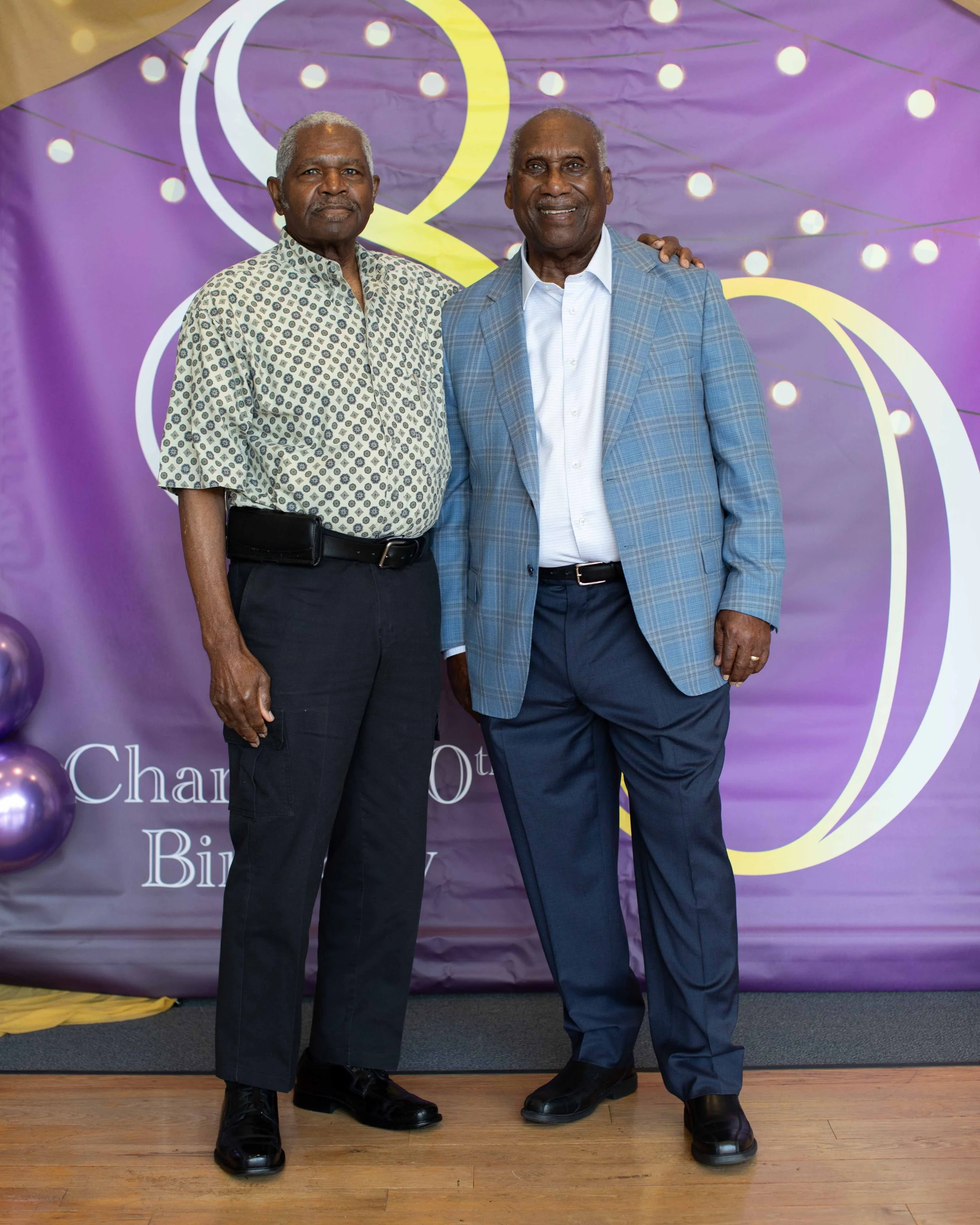 Two older men standing close together at a celebration, one with his arm around the other's shoulder, smiling at the camera. They are dressed in business casual attire with a purple decorated backdrop behind them.