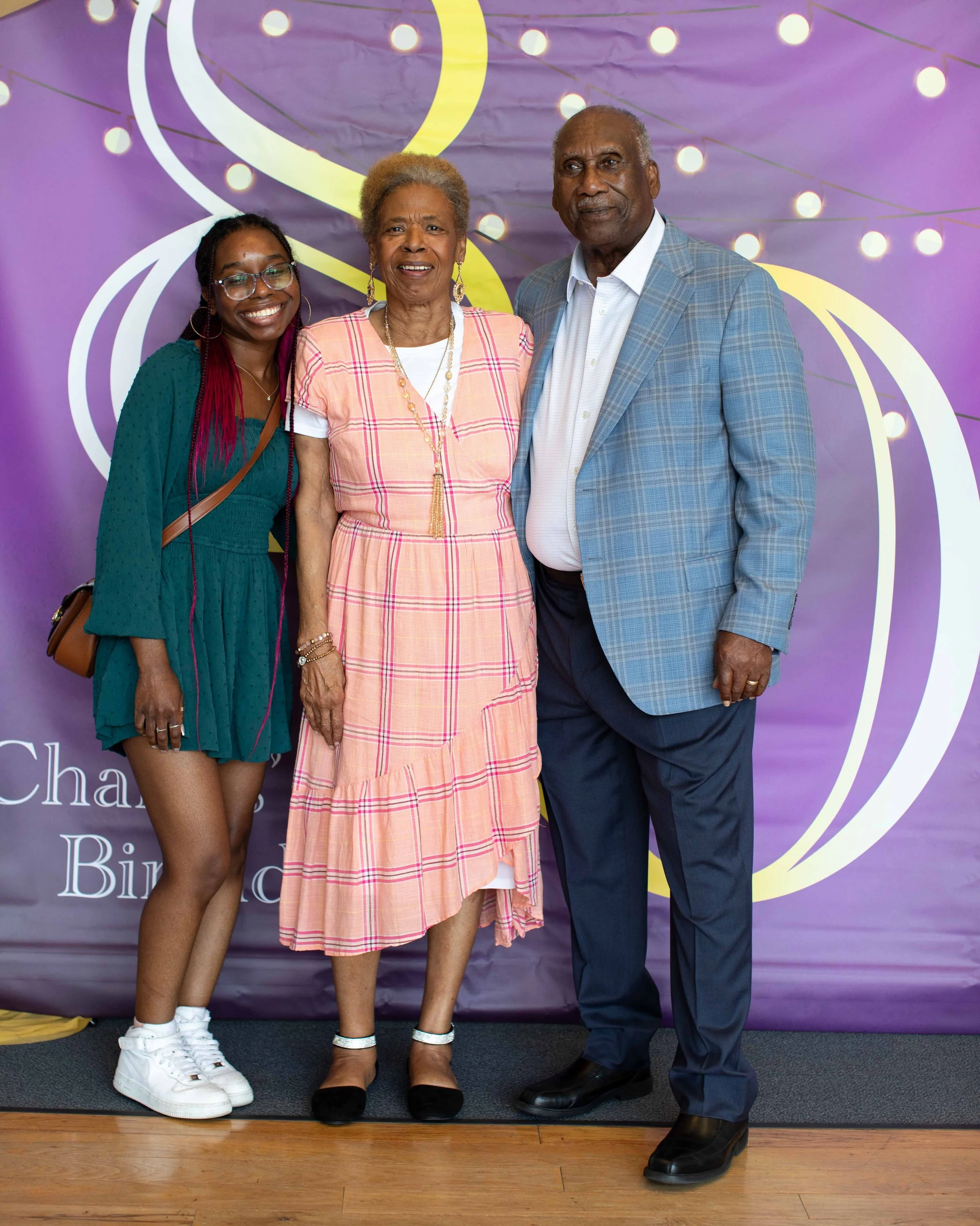 Three people standing together in front of a purple and yellow decorative backdrop. The person on the left is a young woman with dark skin and braids, wearing glasses, a teal dress, and white sneakers. The middle person is an elderly woman with gray 