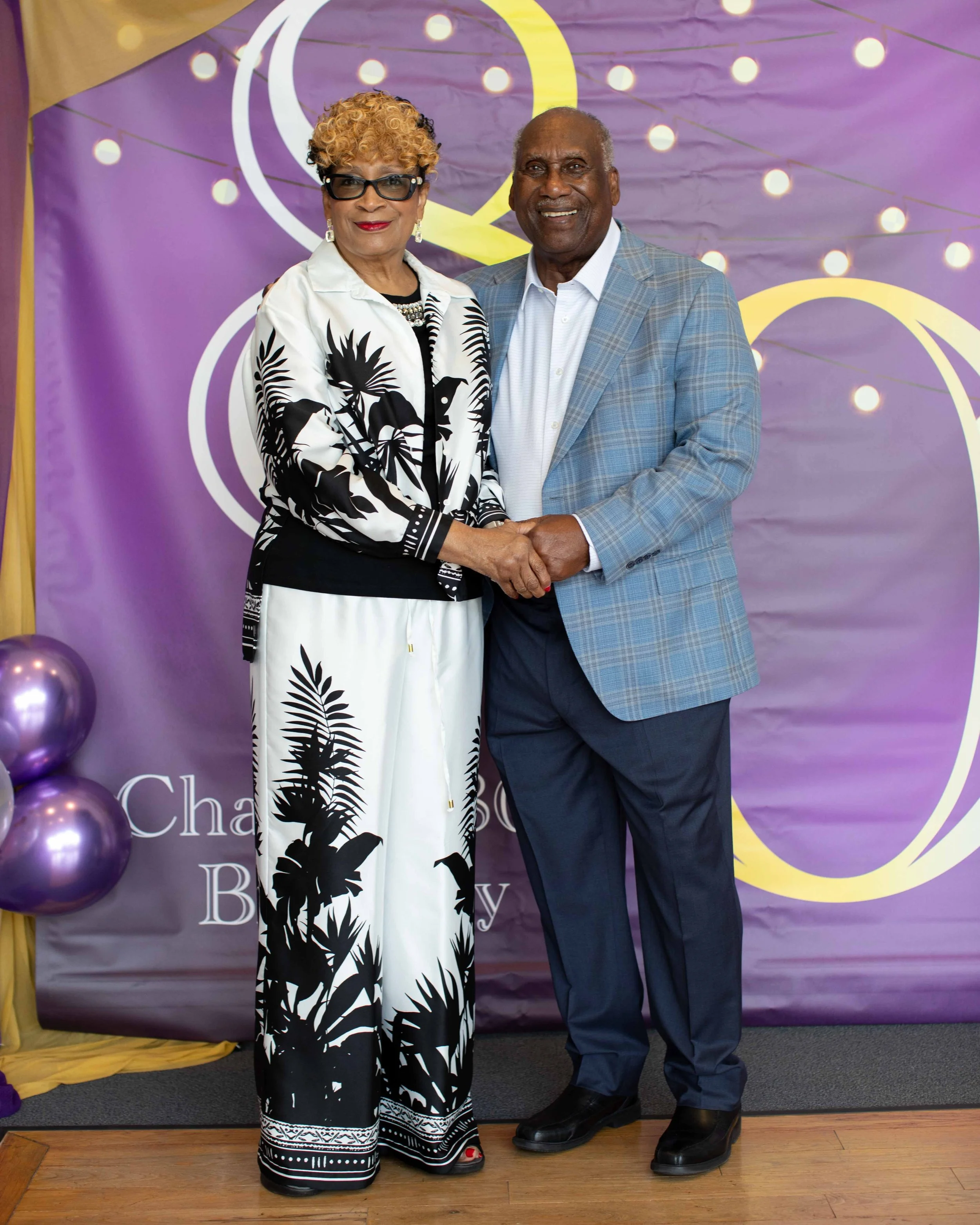 A couple standing in front of a purple backdrop with lights, holding hands and smiling, dressed in formal attire, at an event.