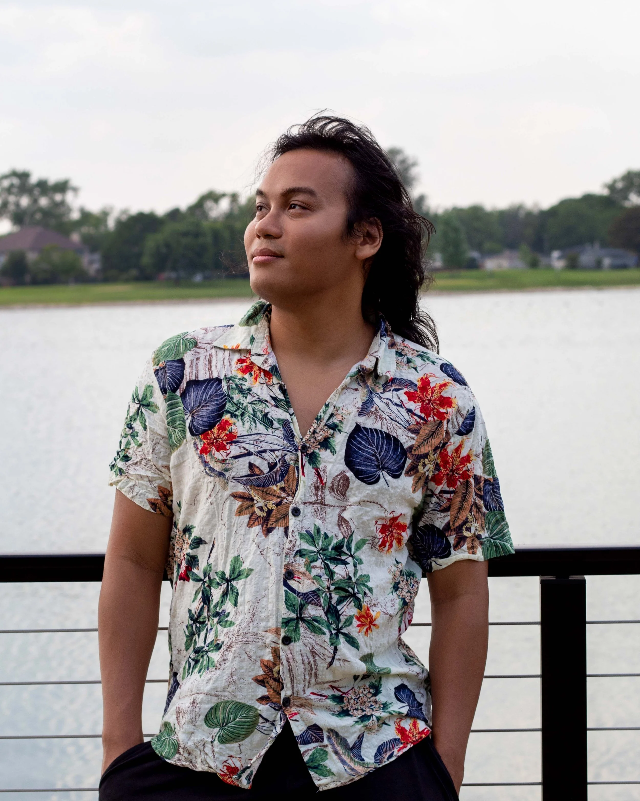 A man with long black hair wearing a colorful tropical patterned short-sleeved shirt stands outdoors by a body of water with trees and houses in the background, looking to the side.