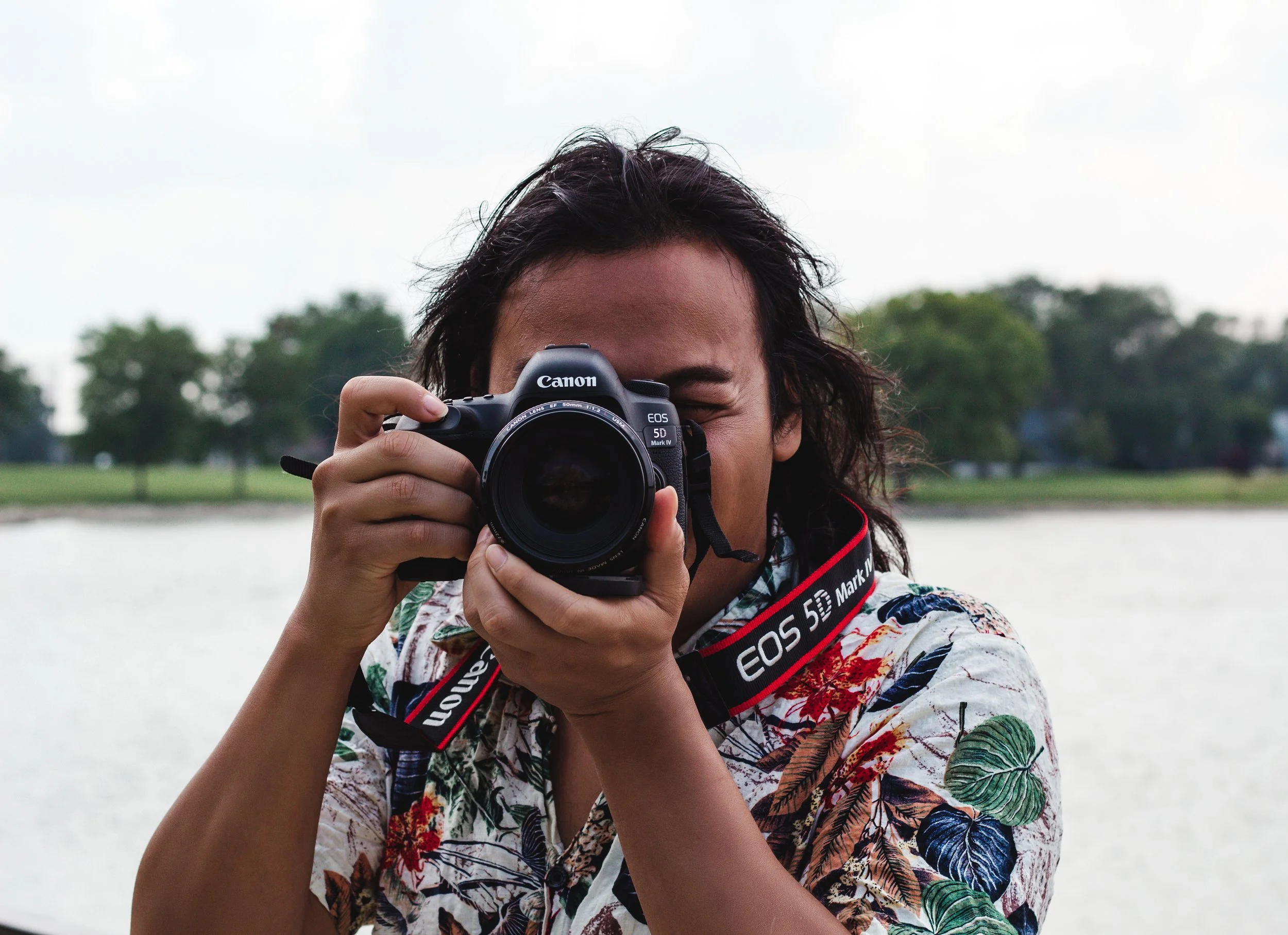 Person with dark hair taking a photo with a Canon EOS 5D Mark IV camera outdoors. They are wearing a floral patterned shirt and standing near a body of water with trees in the background.