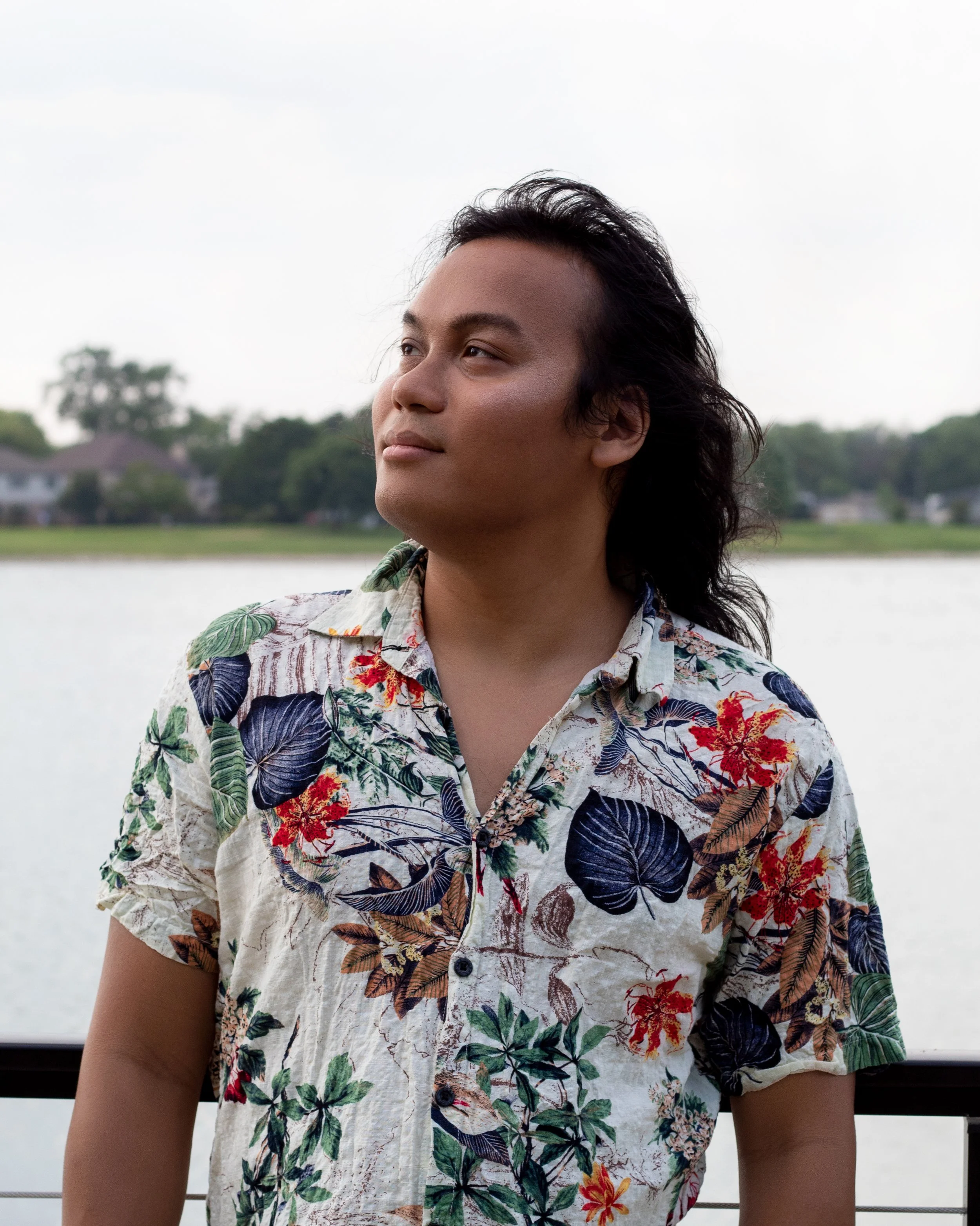 A person with long dark hair hairstyle, wearing a colorful tropical floral shirt, standing outdoors near a body of water with green trees and houses in the background.
