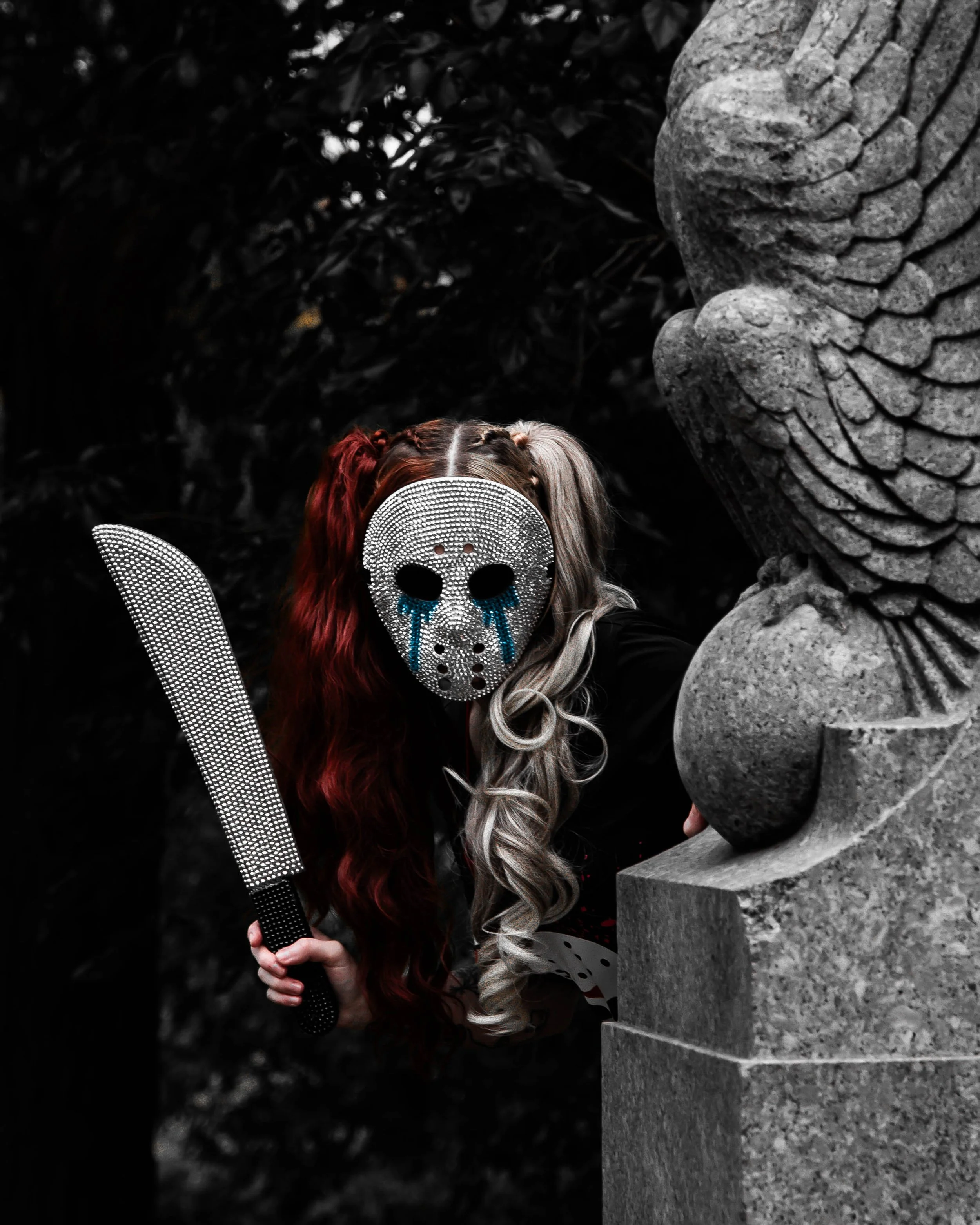 Person with long, curly hair wearing a skull-shaped mask made of small shiny beads, holding a large dagger, standing in front of a stone sculpture in a dark environment.