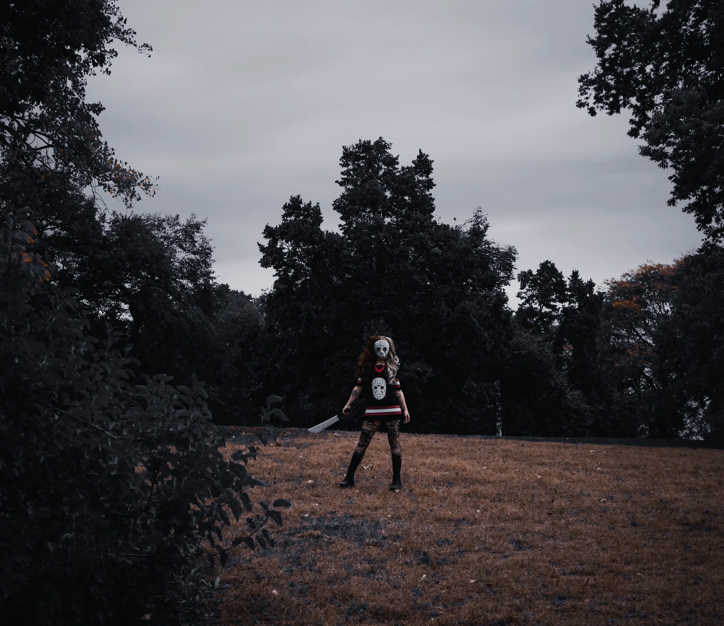 A person dressed in a skeleton Halloween costume with a skull mask, holding a sword, standing outdoors among trees on a cloudy day