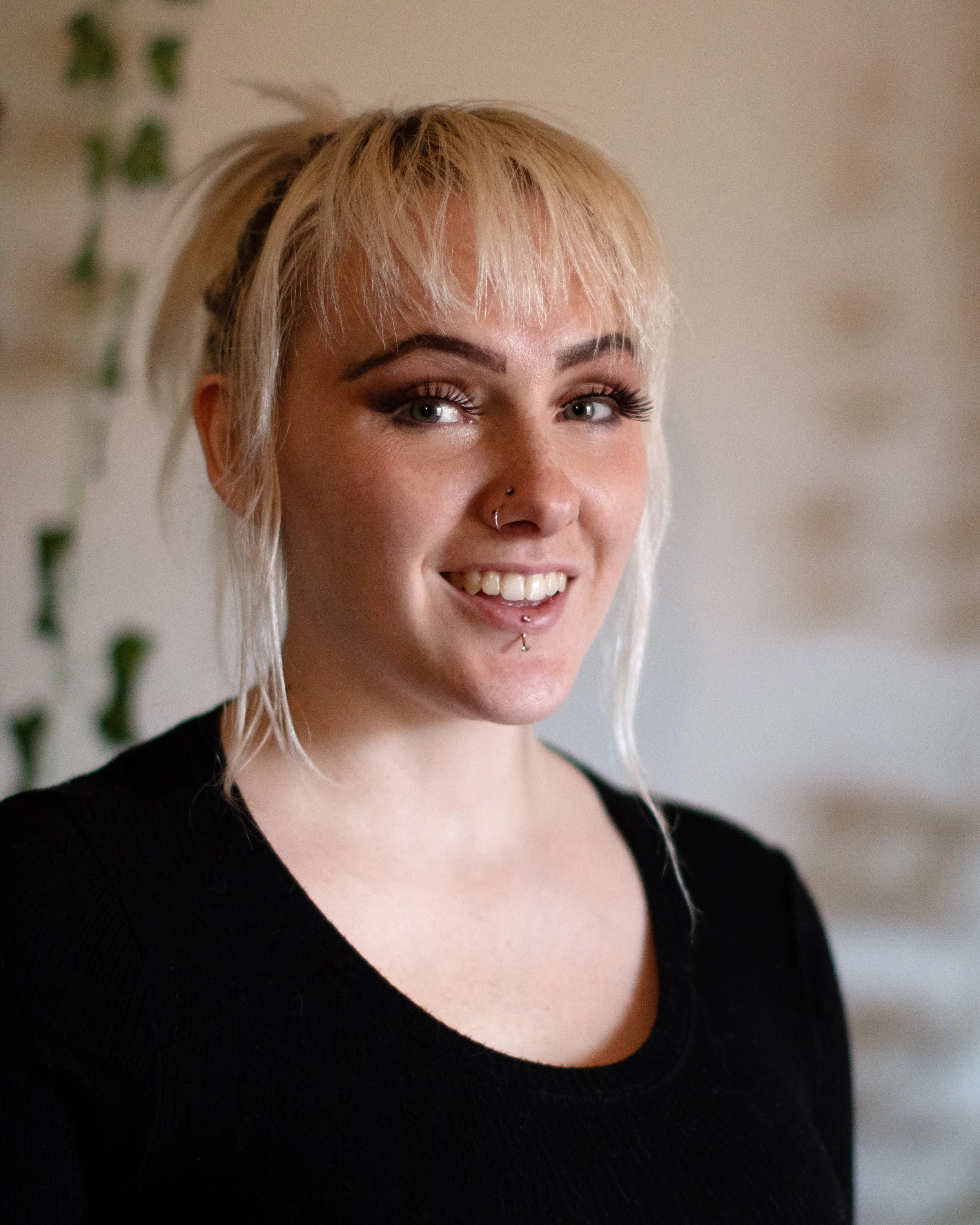 A woman with platinum blonde hair styled in a casual updo, dark eye makeup, and a nose piercing, wearing a black top, smiling softly, in an indoor setting with blurred background.