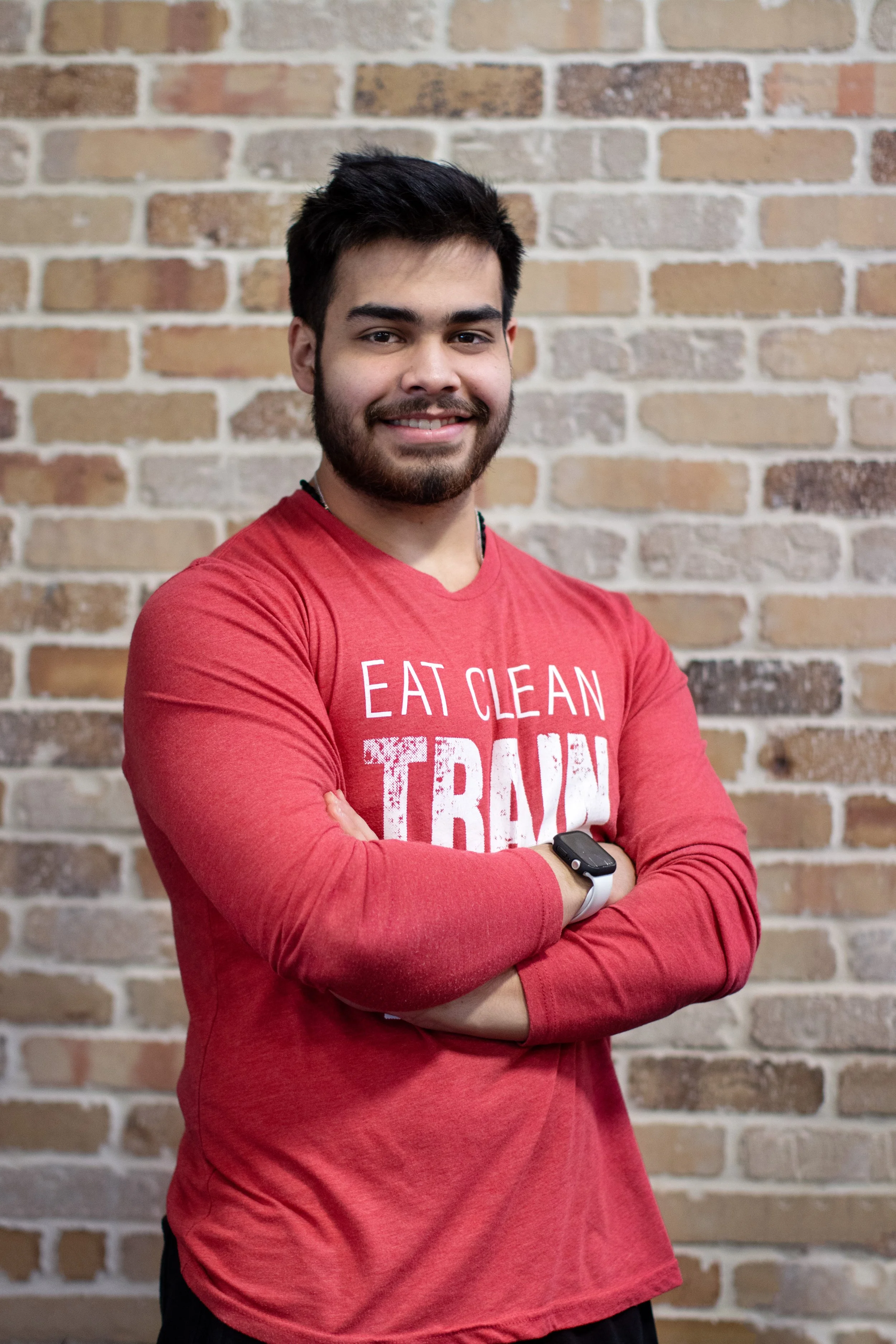 A young man with dark hair and a beard is smiling and standing in front of a brick wall. He is wearing a red long-sleeved shirt with text and has his arms crossed, with a smartwatch on his left wrist.