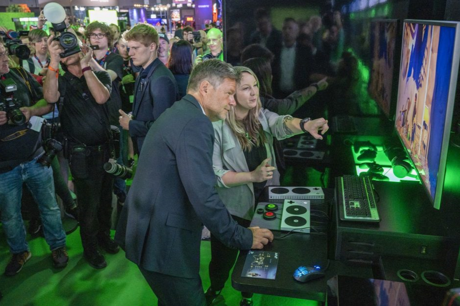 Kaitlyn at a gaming demo station at a conference. The vice chancellor of germany is standing next to her as they both use an adaptive controller. There is a crowd of media with cameras behind them.