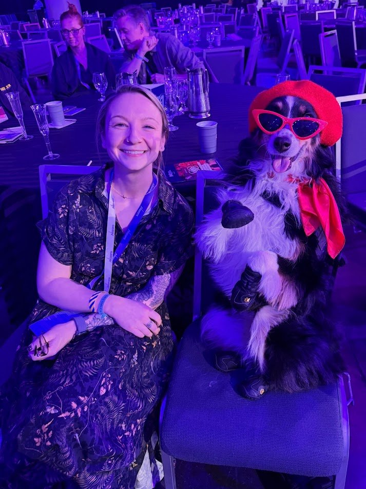 Kaitlyn posing with a australian shepherd service dog. The dog is wearing a red Baret, red cateye sunglasses, a red scarf and is posing with one paw up in the air like she is waving.
