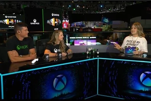 Kaitlyn sitting at a broadcast desk with the Xbox logo on it speaking with the primary "reporter" on the other side of the desk