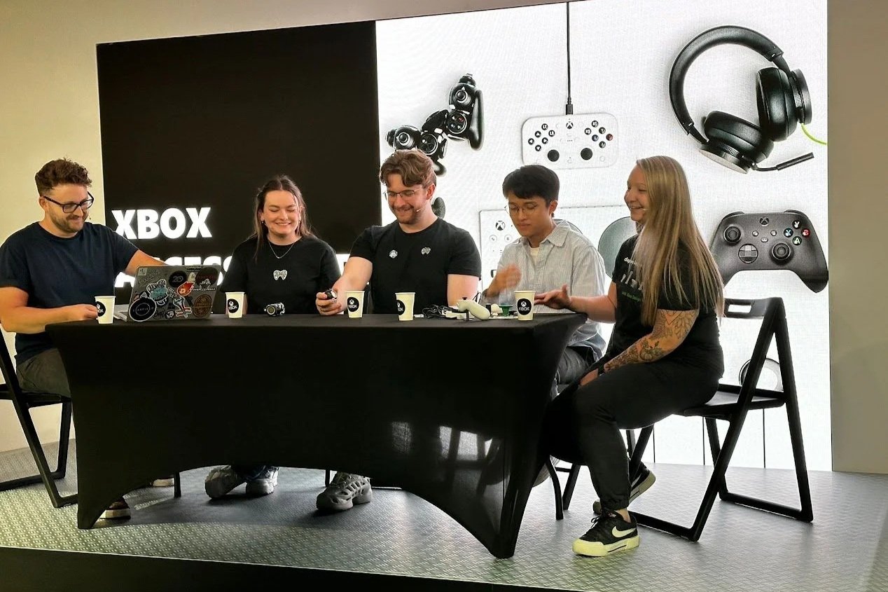 Kaitlyn sitting at a desk on a stage alongside 5 other people. They are smiling as they talk about adaptive controllers to the crowd.