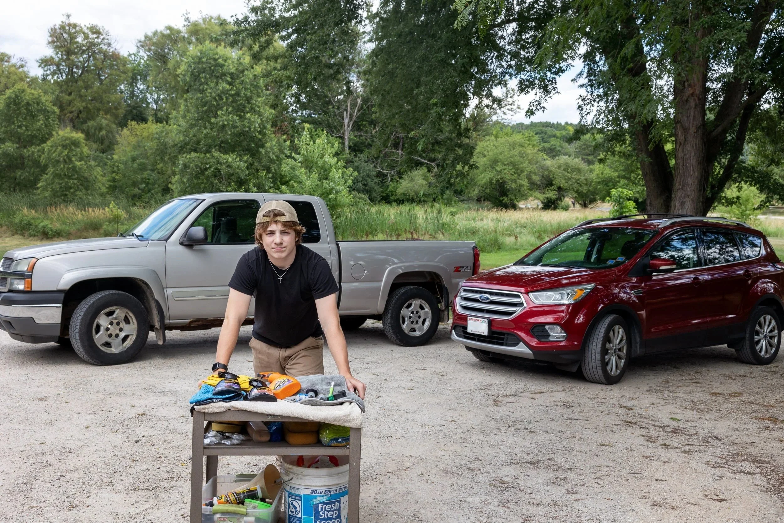 A young boy with a beige cap and black t-shirt crouching next to a cart filled with tools and supplies, in a parking lot with two cars and a background of trees and greenery.