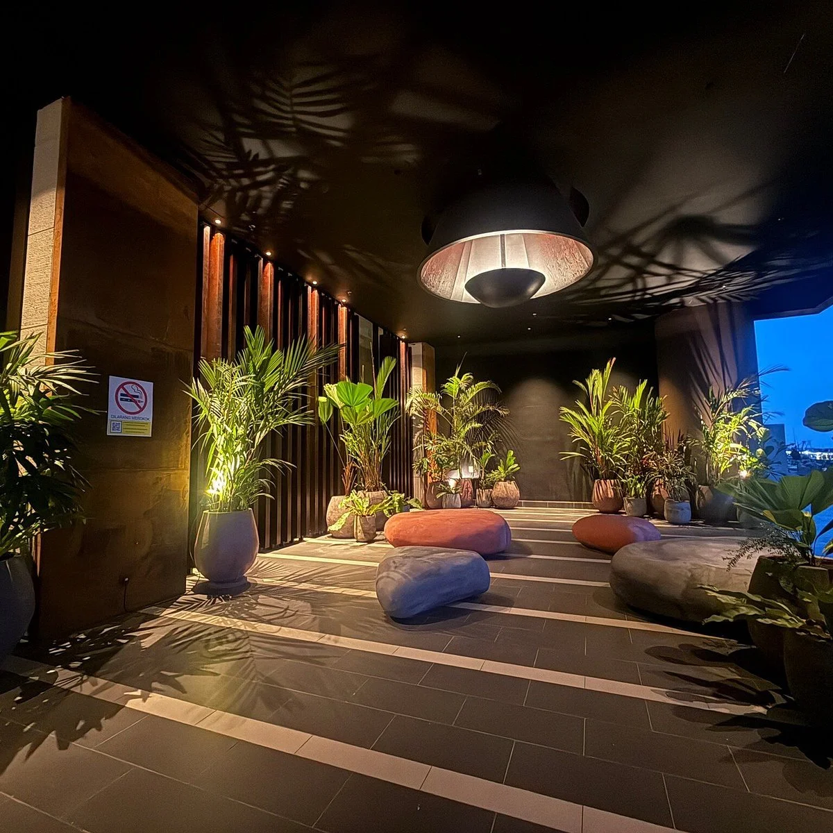 Nighttime outdoor lounge area with potted tropical plants, large decorative rocks, and modern seating, illuminated by warm lighting and a large overhead lamp, casting shadows on the tiled floor.