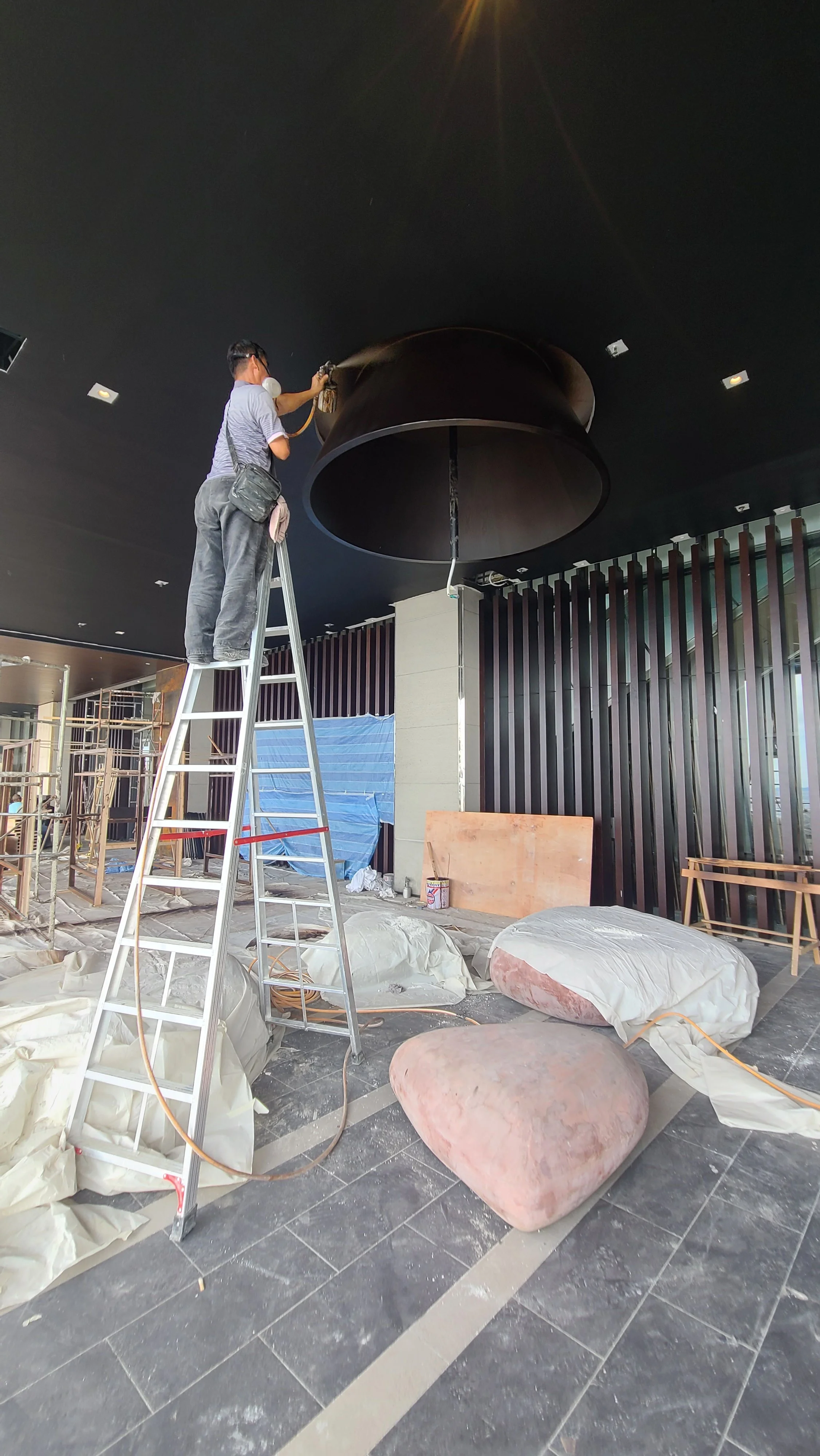 A person standing on a ladder, polishing a large black interior ceiling fixture. The scene appears to be in the process of construction or renovation, with protective coverings, construction materials, and tools scattered on the floor.