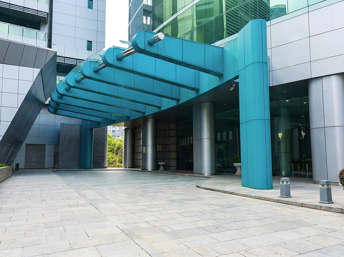 Modern glass and steel building entrance with a blue awning, metallic pillars, and paved sidewalk.