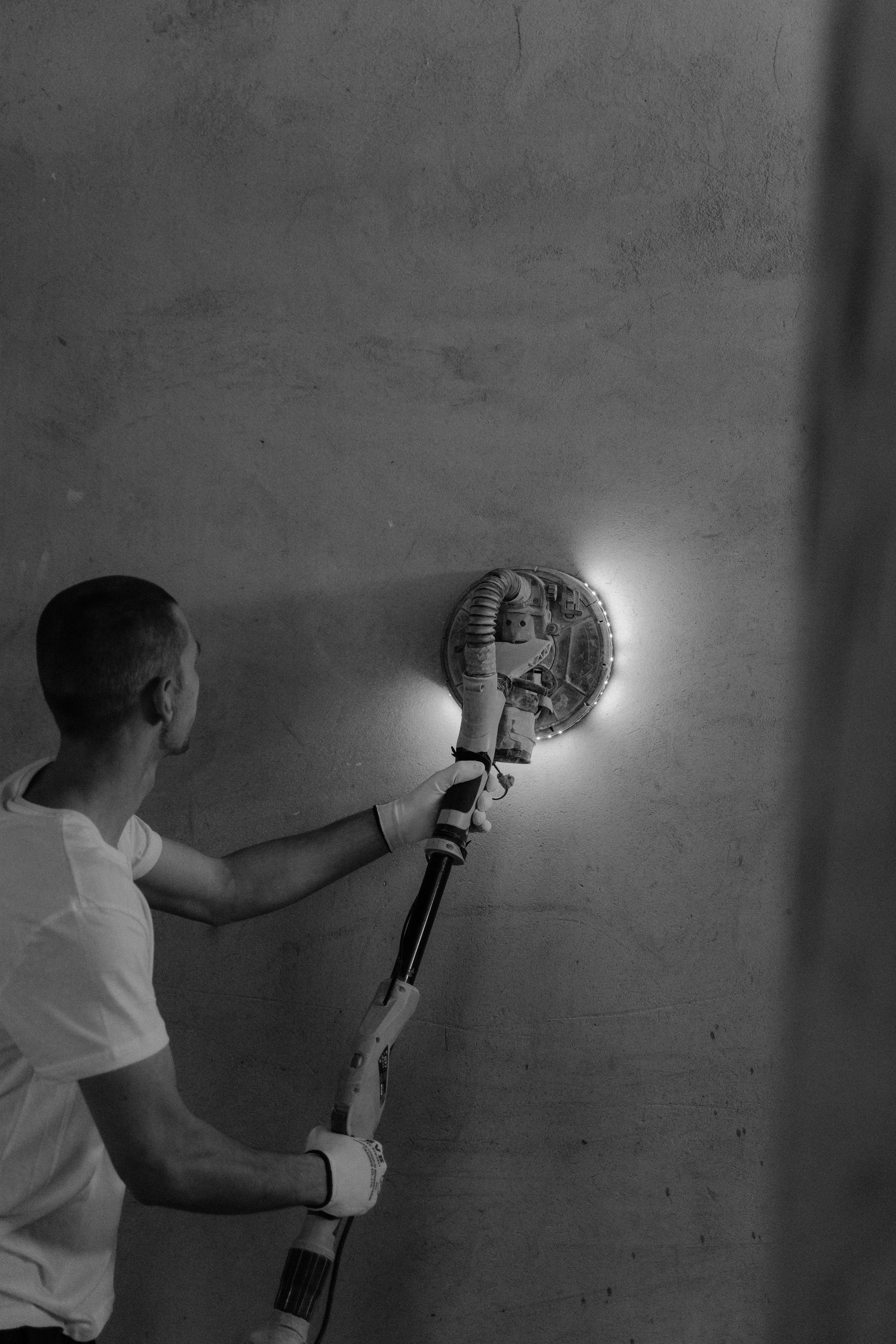 A man in a white t-shirt is using a power drill to work on a round hole in a wall. The drill is connected to a vacuum hose, and the man is wearing gloves. The background is a plain wall.