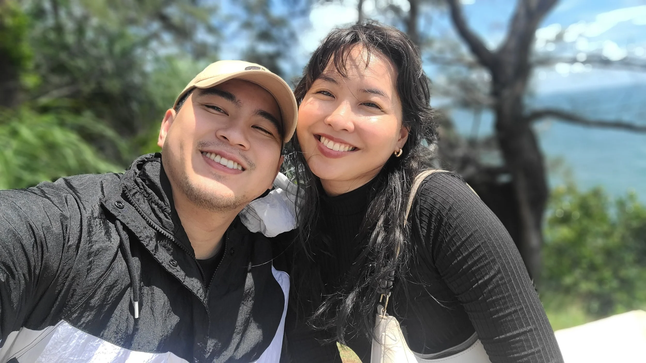 Two people taking a selfie outdoors with a large tree and blue sky in the background. One person is wearing a beige cap and black jacket, and the other person has dark hair and is smiling.