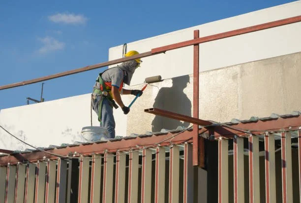 A construction worker painting a wall on a building's rooftop with a roller brush.