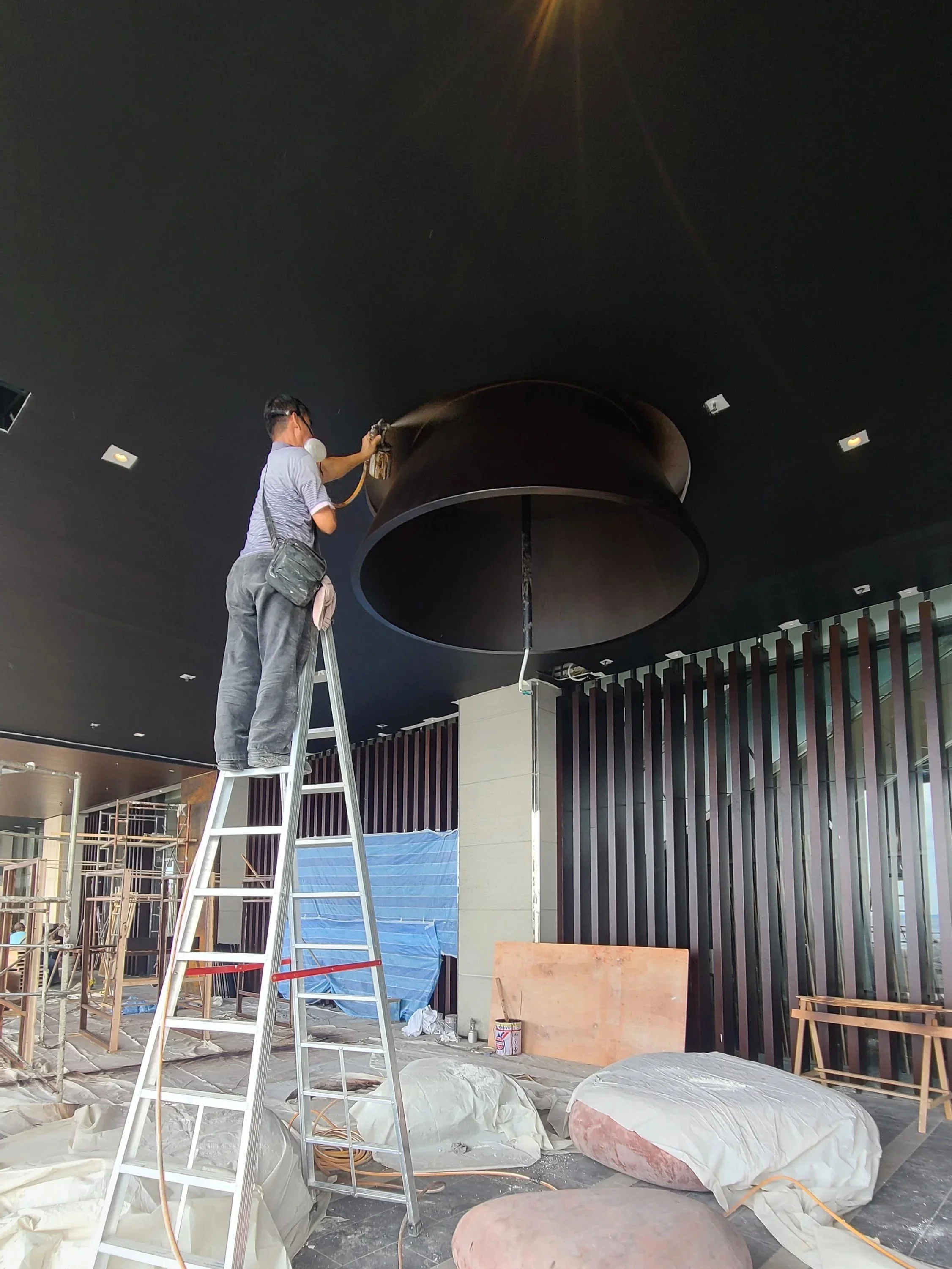A person standing on a ladder cleaning or inspecting a large circular structure hanging from a ceiling inside a building under construction or renovation. The room has construction materials, tools, and protective coverings on the floor.