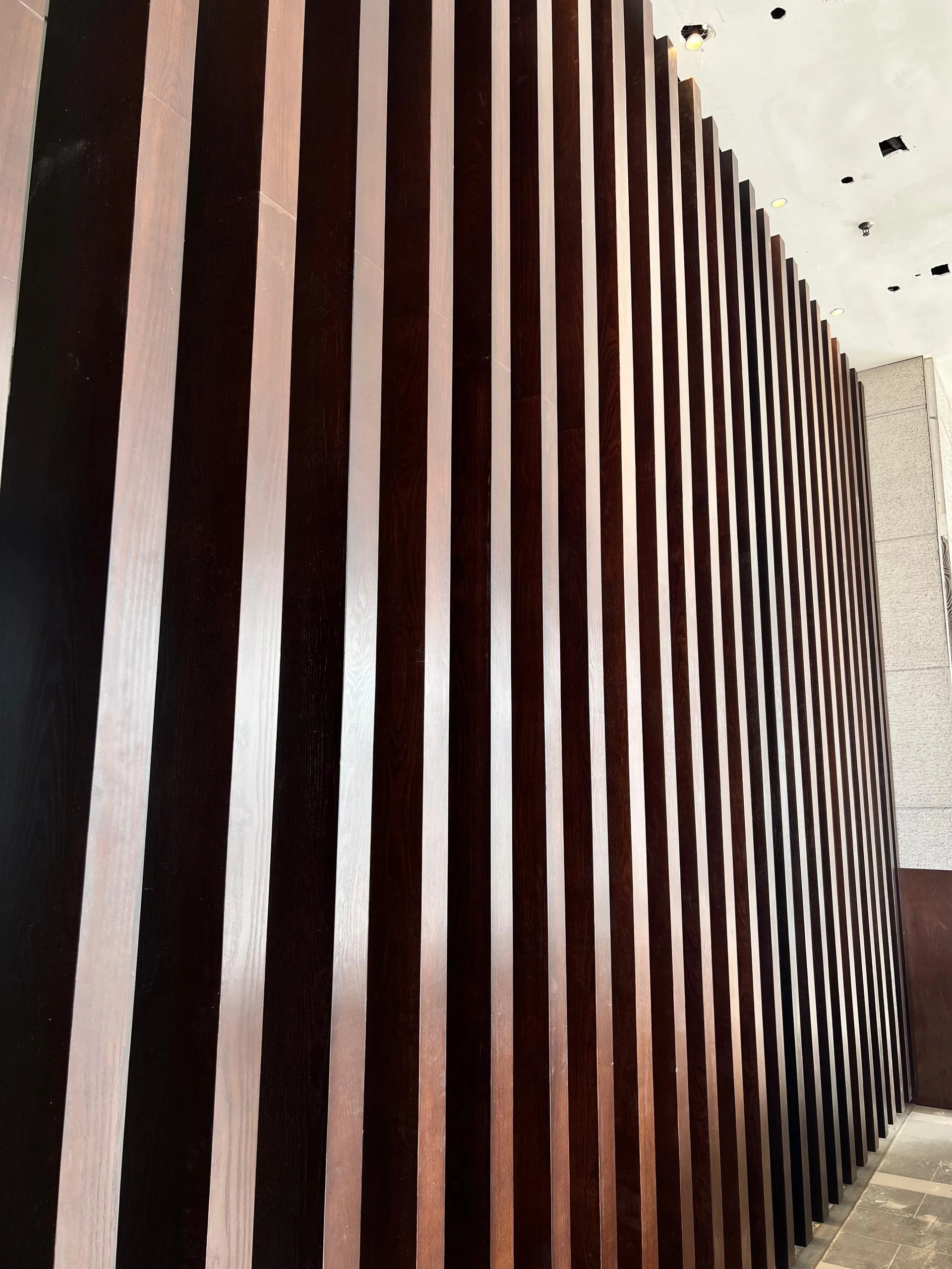 Interior wall with evenly spaced vertical wooden slats of alternating dark and light brown shades in a modern design.