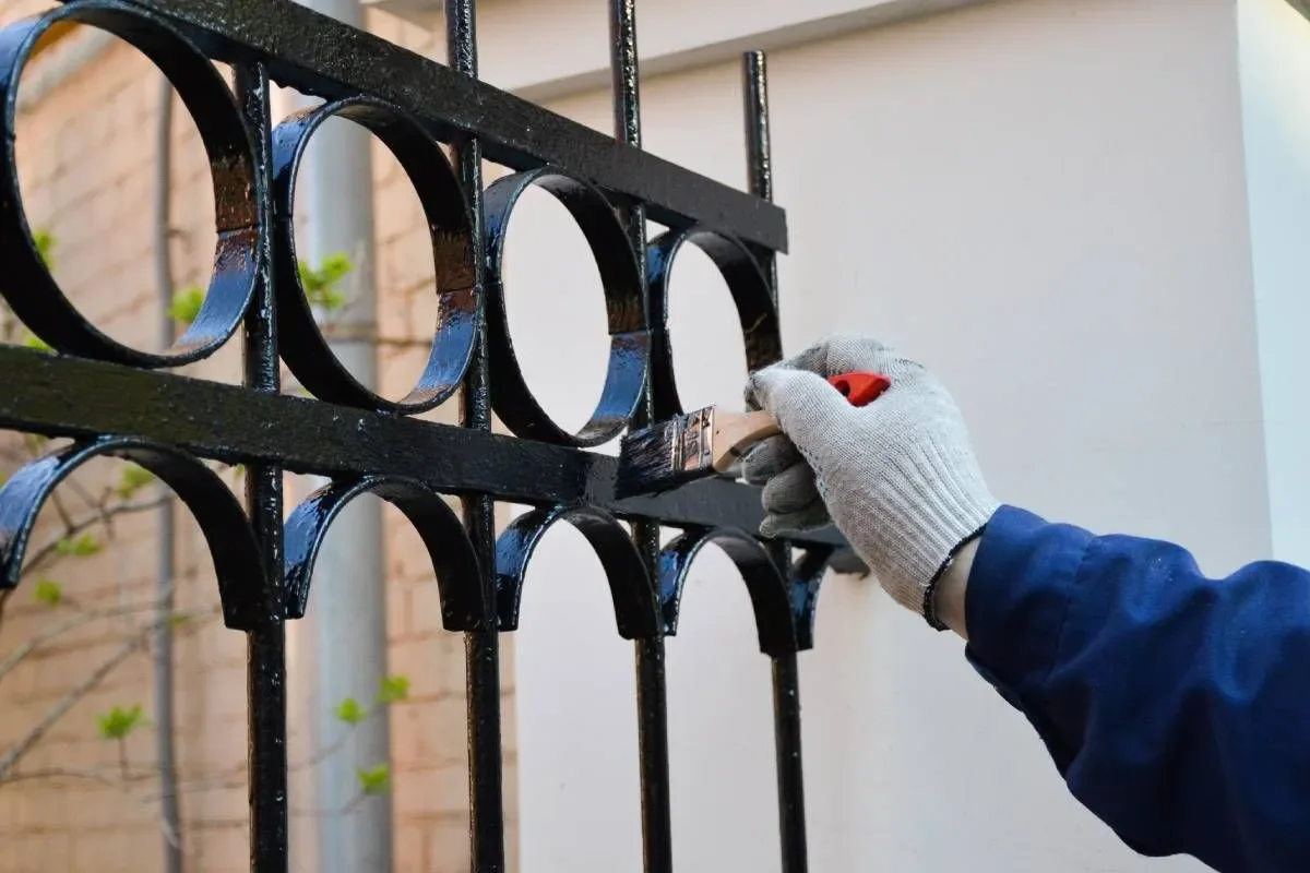 A person wearing work gloves is painting or touching up a black metal fence with a small brush.