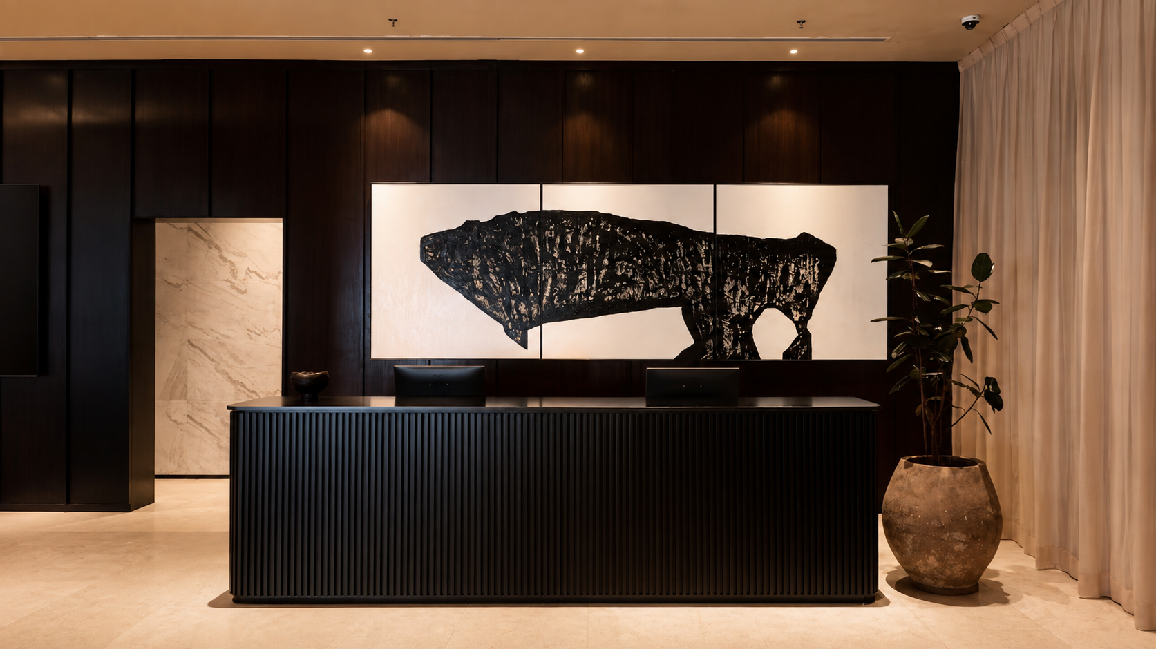 Modern reception desk in a hotel lobby with a large black and white artwork of a bear on the wall behind it. Includes a potted plant to the right of the desk and curtains on the far right.