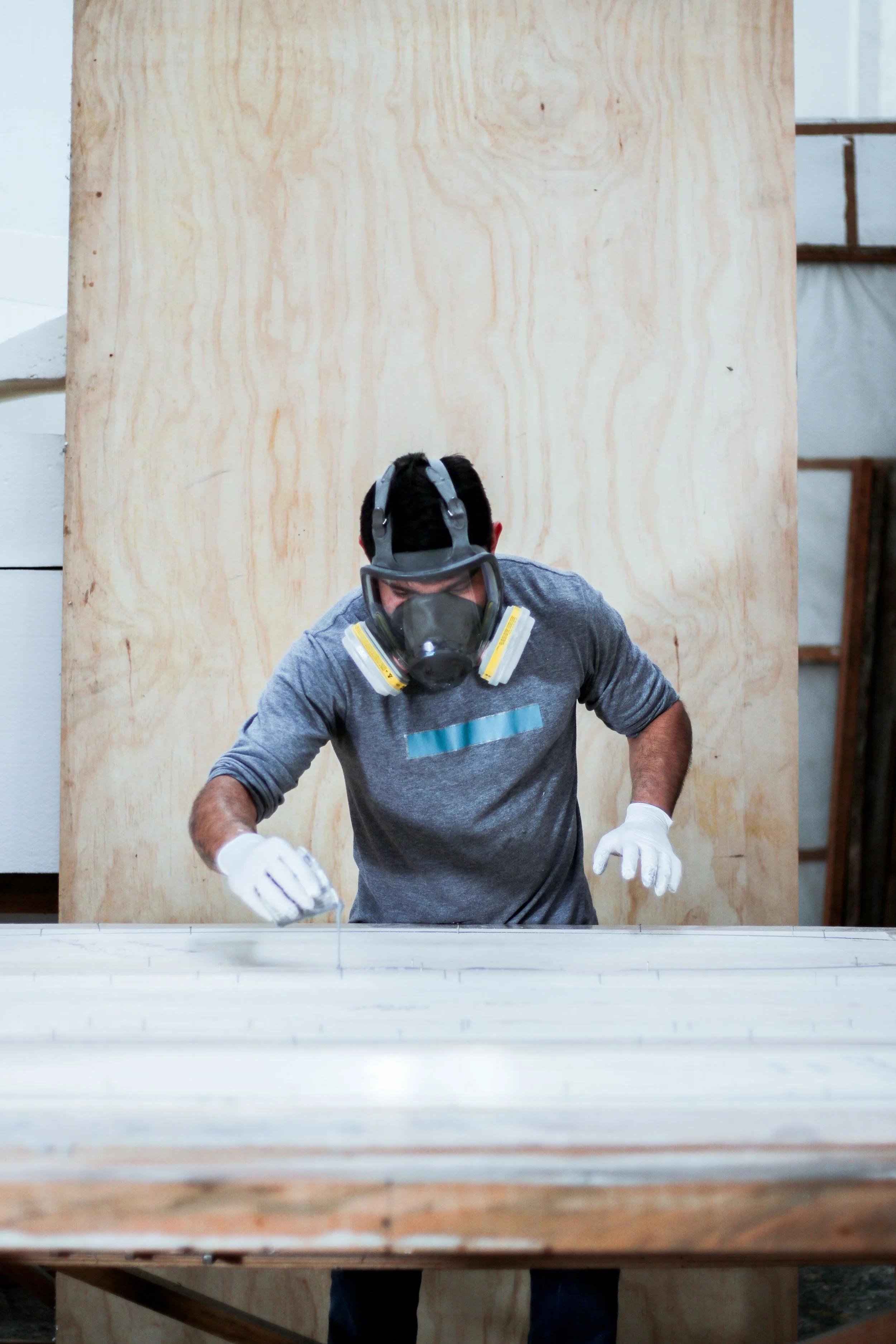 A man wearing a respirator mask, gloves, and a gray shirt is working on a project behind a large wooden panel in a workshop.