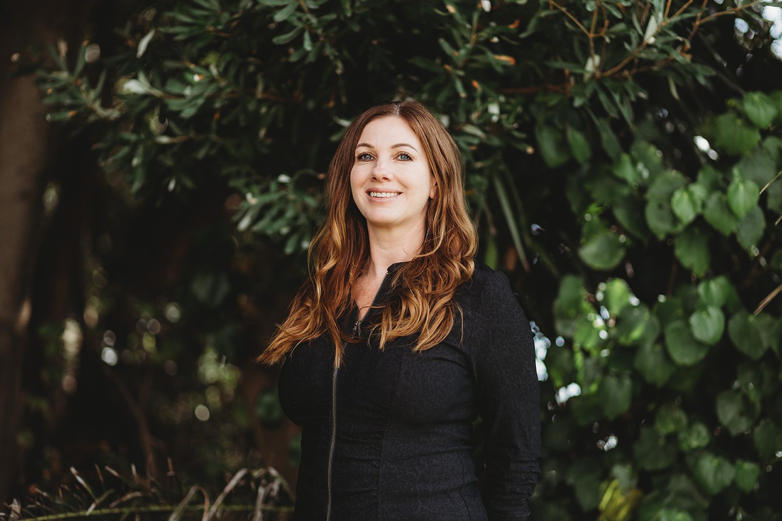 A woman with long wavy red hair smiling outdoors in front of green leafy bushes.