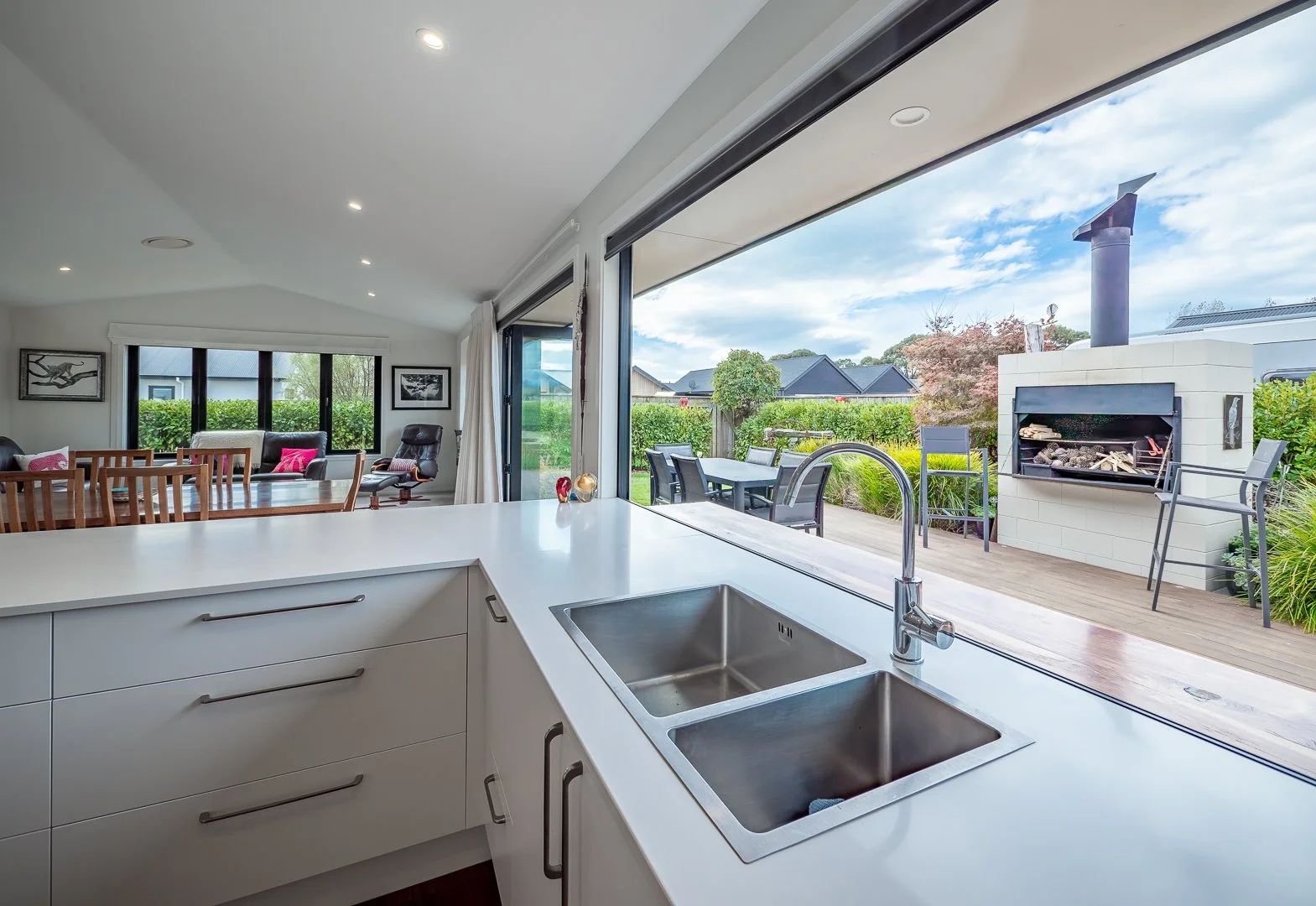 Modern kitchen with white countertop, stainless steel sink, open view to outdoor patio with seating, and fireplace in backyard.
