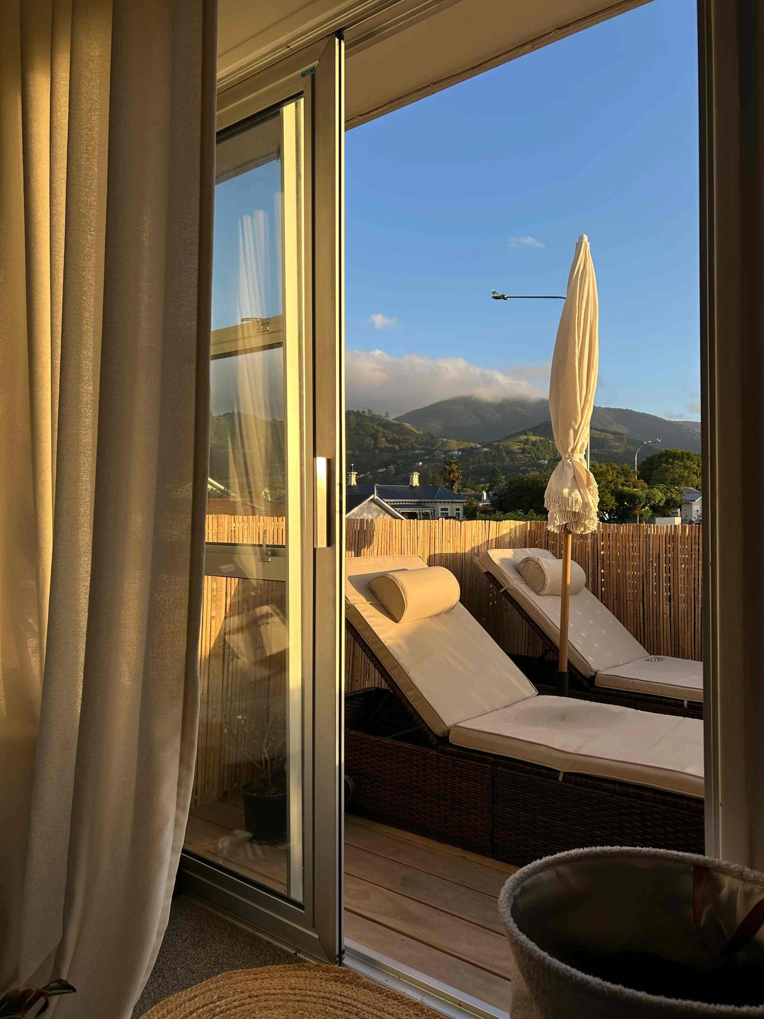 View through sliding glass door showing outdoor patio with two lounge chairs, an umbrella, mountain scenery in the background, and a blue sky with some clouds.