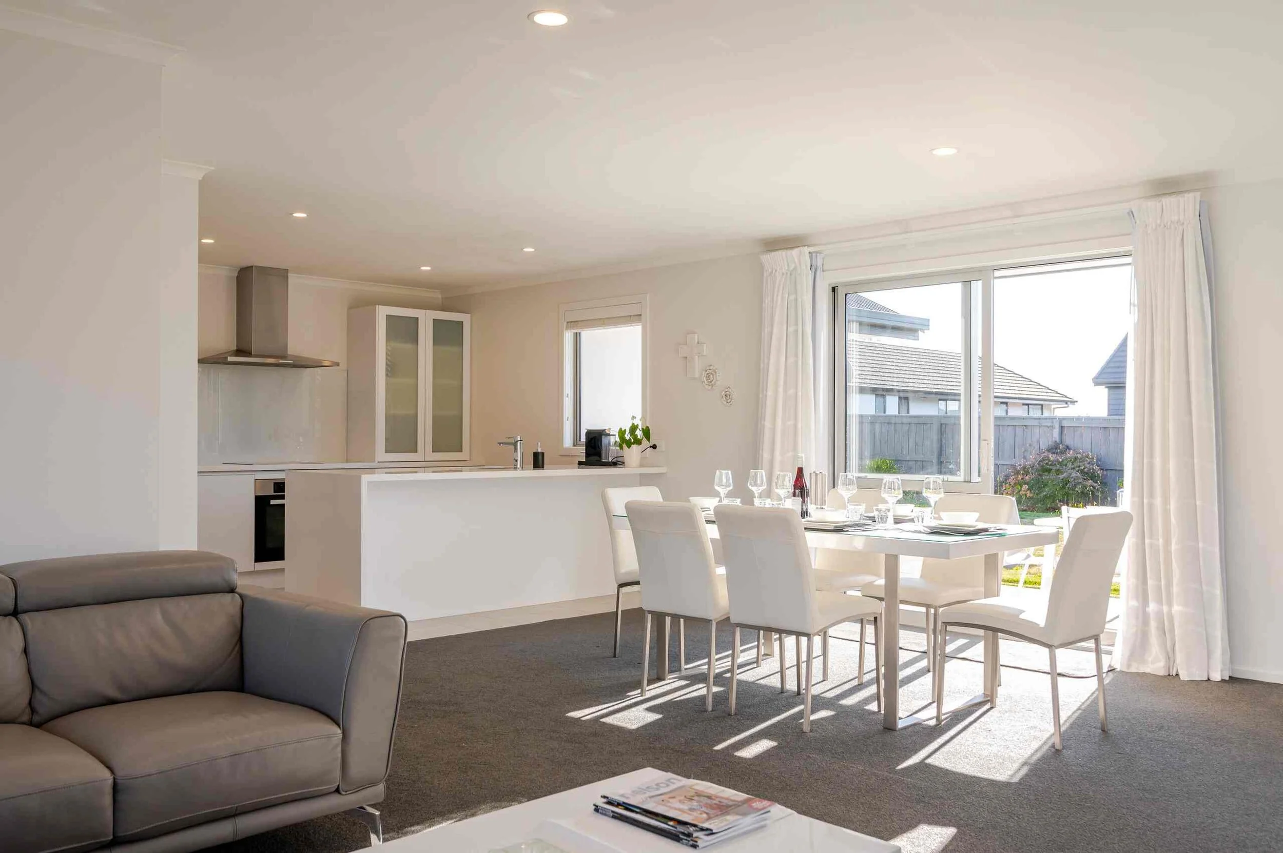 Bright living room with a grey sofa, white dining table set with six chairs, large windows with white curtains, a kitchen area with white cabinetry, and a view of neighboring houses outside.
