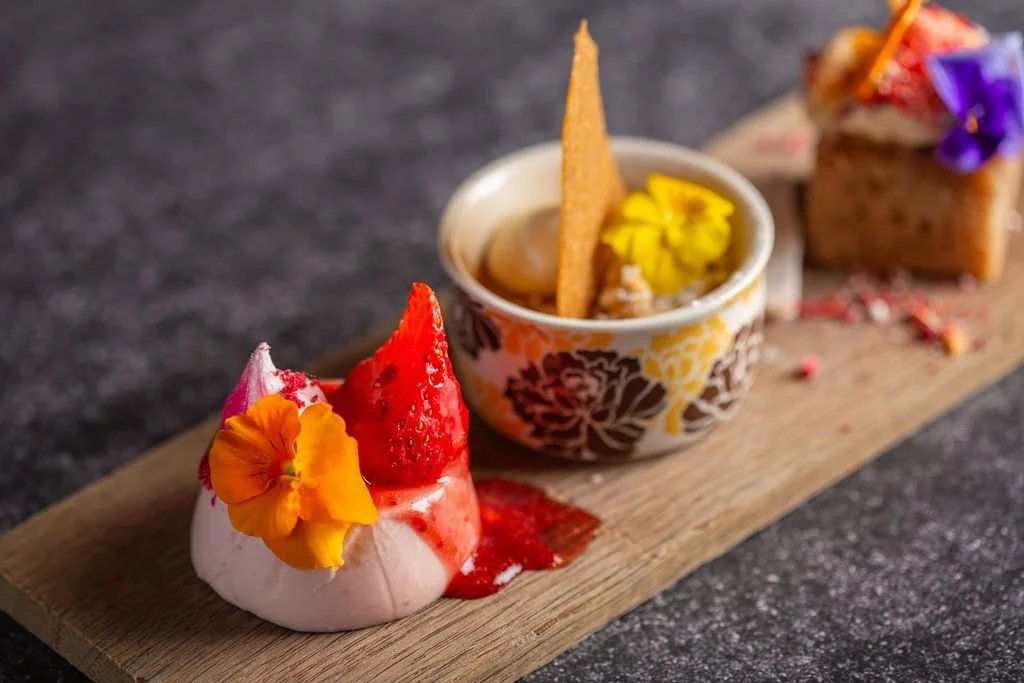 Dessert platter with meringue topped with strawberry and edible flower, a small bowl of ice cream with a cookie and flower decoration, and a piece of cake with floral decoration on wooden board.