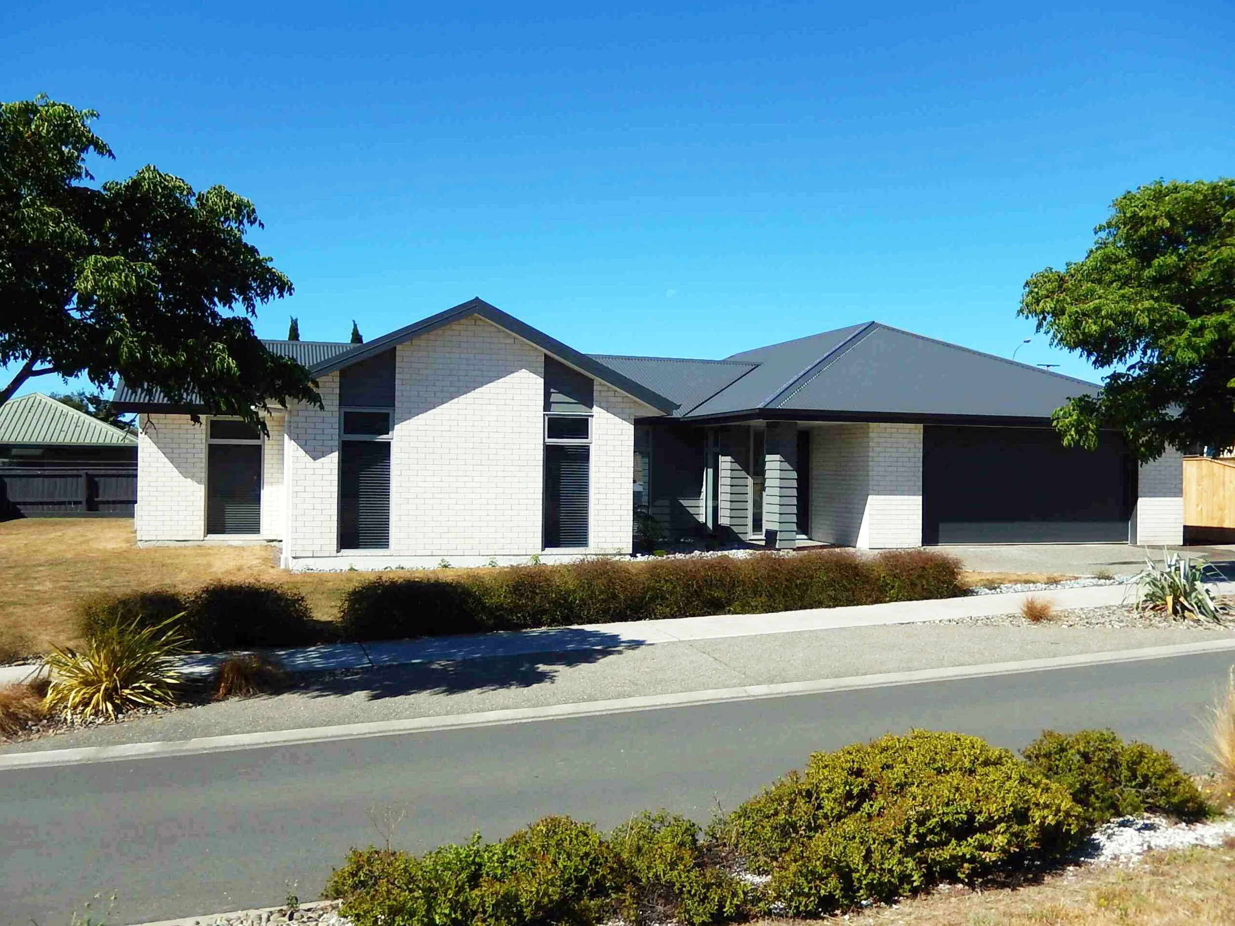 A modern white brick house with a dark gray roof, surrounded by trees and low shrubs, located on a residential street under a clear blue sky.