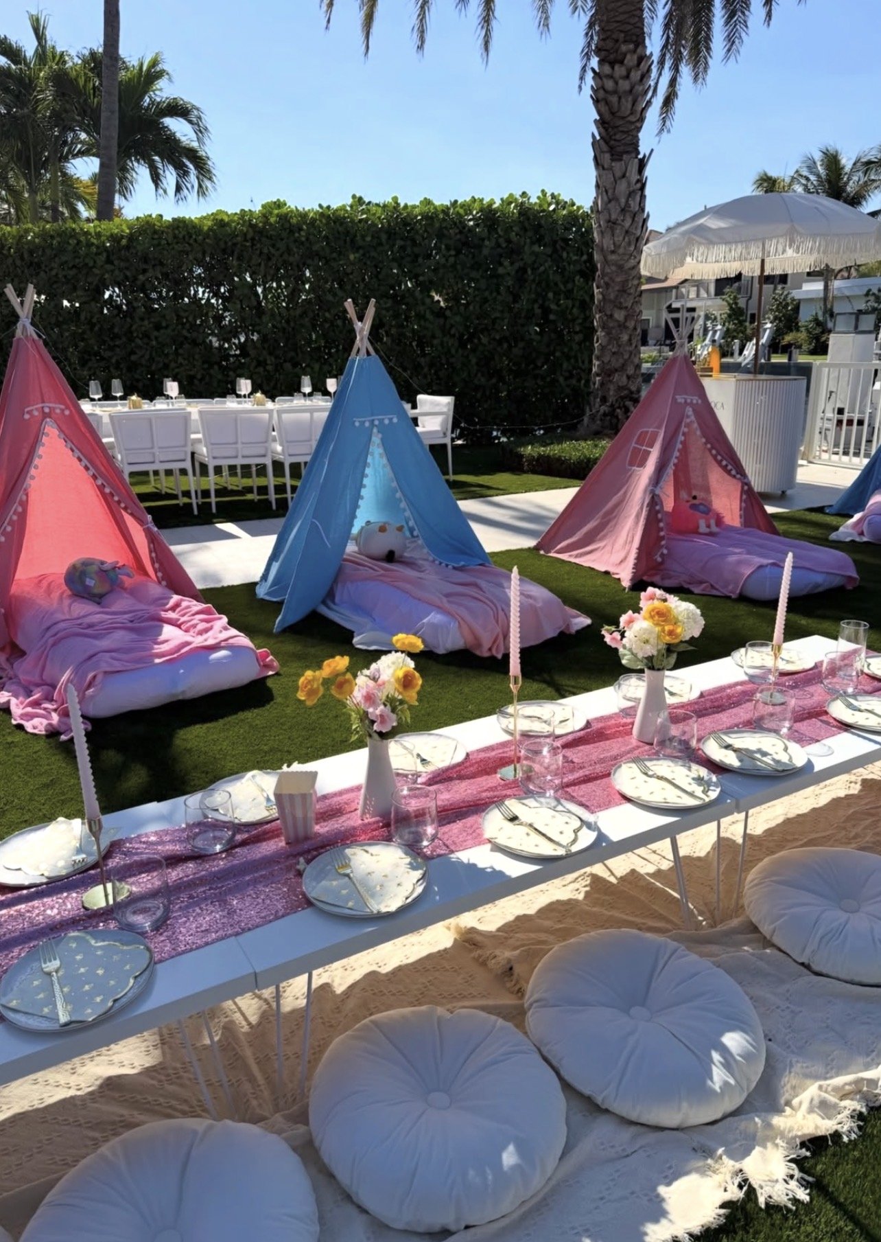 Outdoor party setup with pink and blue teepees on grass, decorated with plush toys. In front, a long table with a pink table runner, plates, glasses, cutlery, and small flower arrangements. Cushions are placed around the table on a sandy surface. Tall palm trees and a hedge provide a tropical backdrop, with clear blue sky overhead.